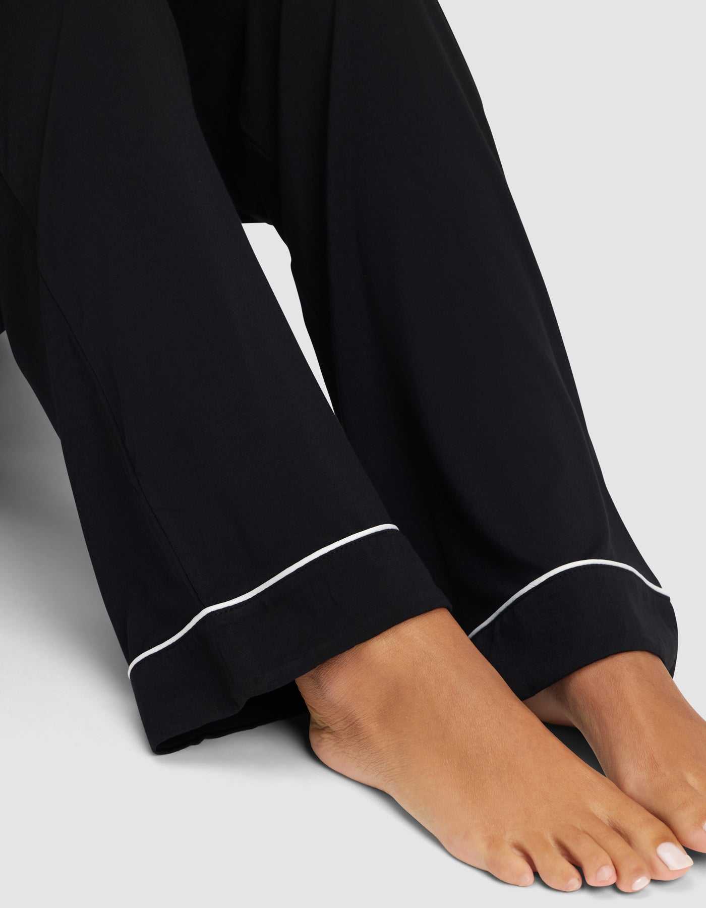 A close-up of bare feet and lower legs in Cozy Earth’s Women's Bamboo Stretch-Knit Long Sleeve Pajama Set, featuring loose black pants with white piping at the hem, set against a plain light gray background. |Color:Black