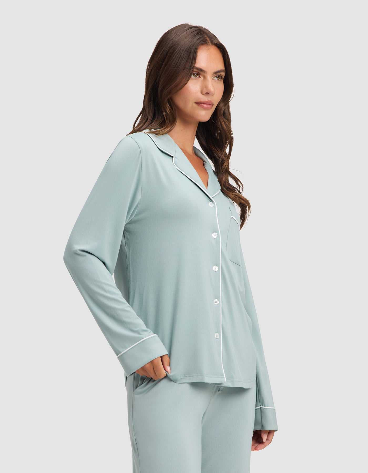 A woman models the Cozy Earth Women's Bamboo Stretch-Knit Long Sleeve Pajama Set in light blue with white piping, one hand in her pocket, standing against a plain light background. |Color:Brook