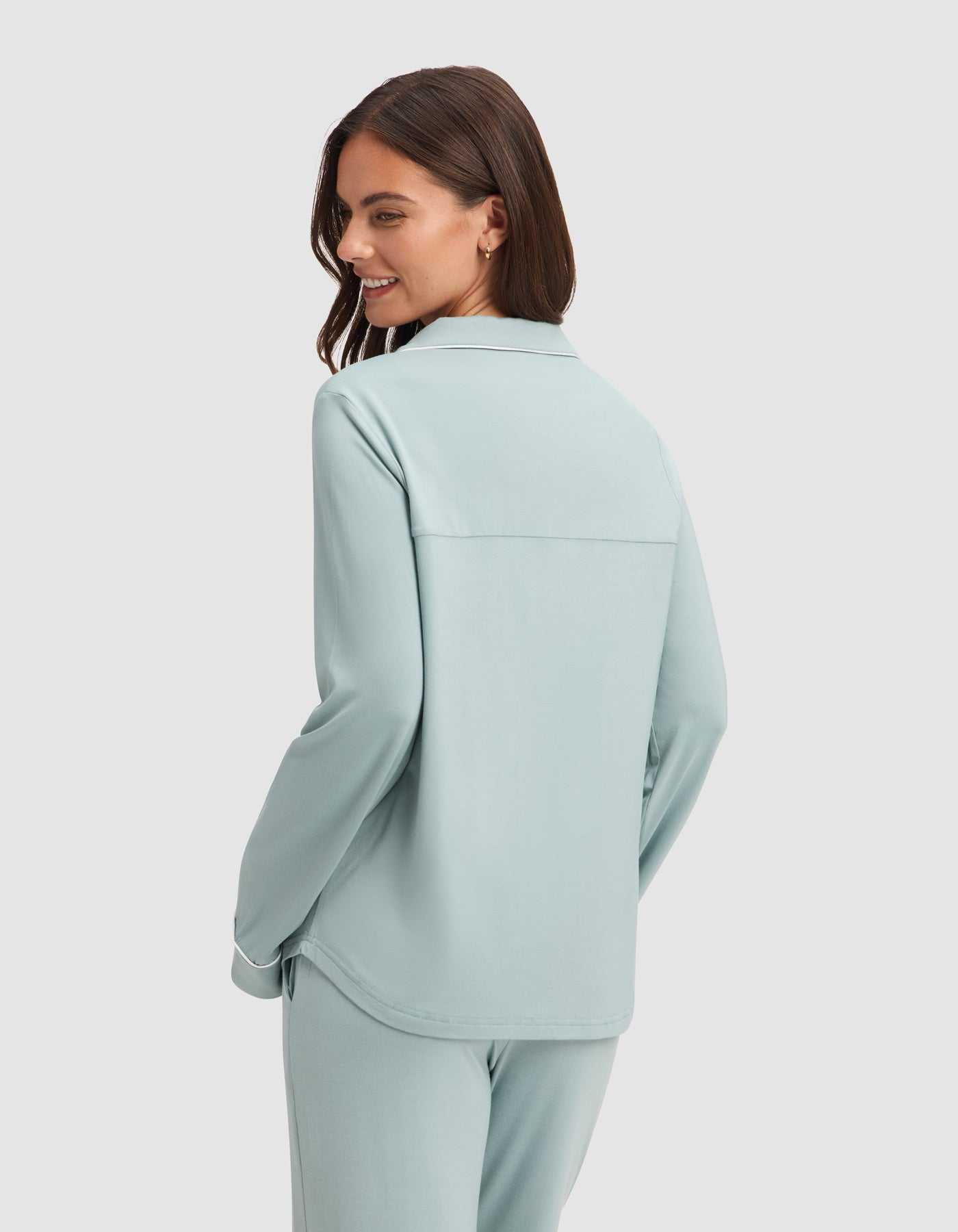 A woman with long brown hair wears the Cozy Earth Women's Bamboo Stretch-Knit Long Sleeve Pajama Set in light blue, standing against a plain white background and smiling over her shoulder. |Color:Brook