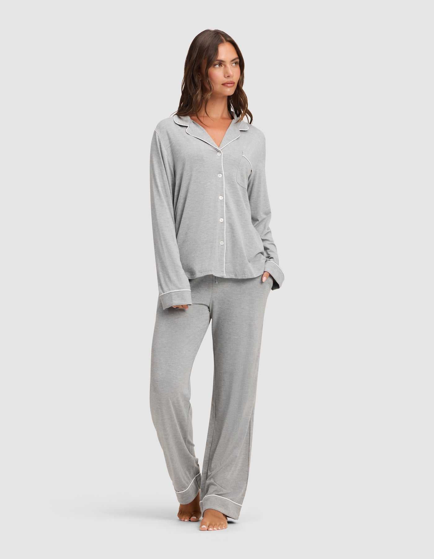 A woman stands barefoot against a plain light background, wearing the Cozy Earth Women's Bamboo Stretch-Knit Long Sleeve Pajama Set in light grey with white piping, featuring a button-up top and matching pants. |Color:Grey
