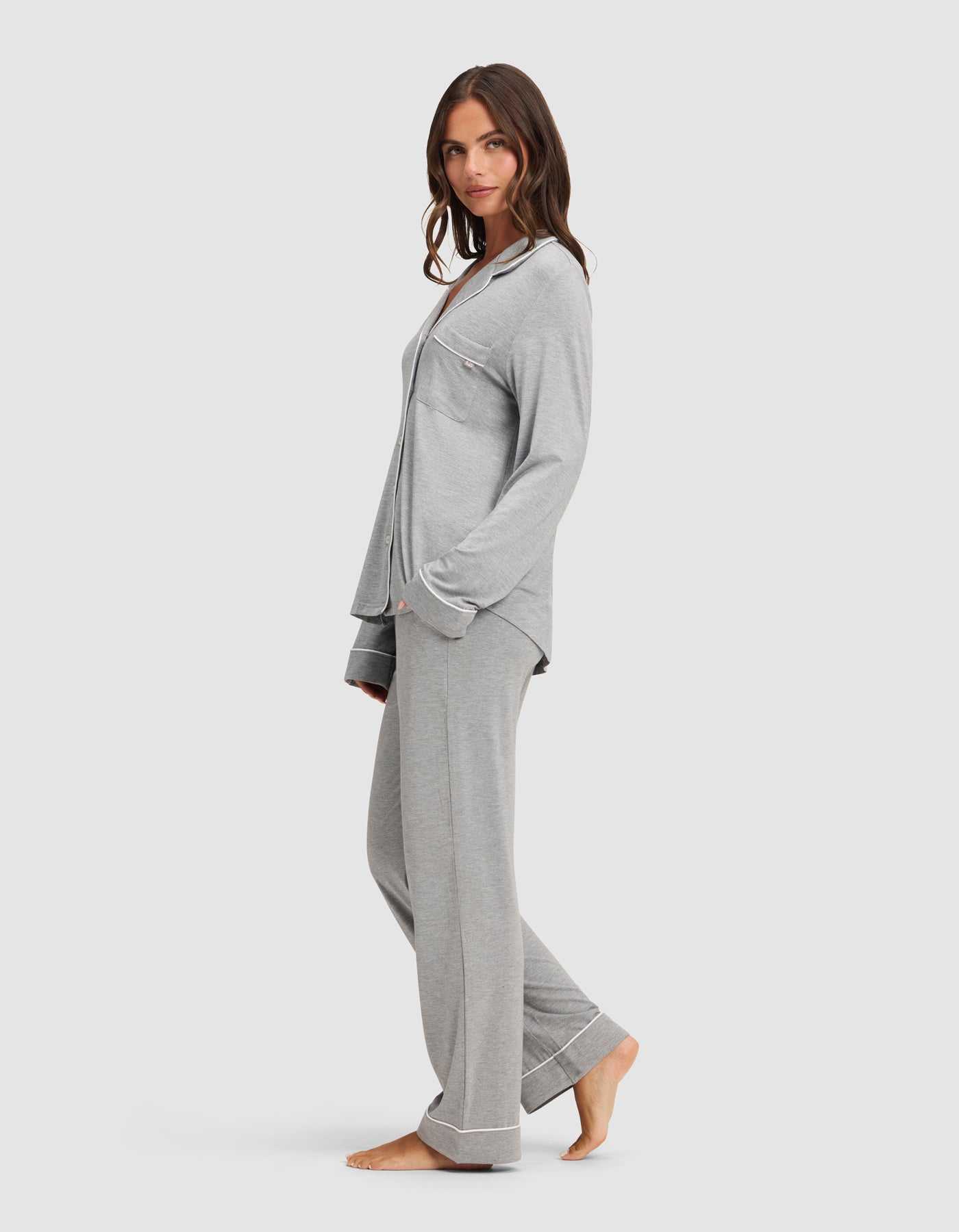 A woman stands barefoot in a relaxed pose, wearing the Cozy Earth Women's Bamboo Stretch-Knit Long Sleeve Pajama Set in light gray with button-up top and chest pocket, against a plain white background. |Color:Grey