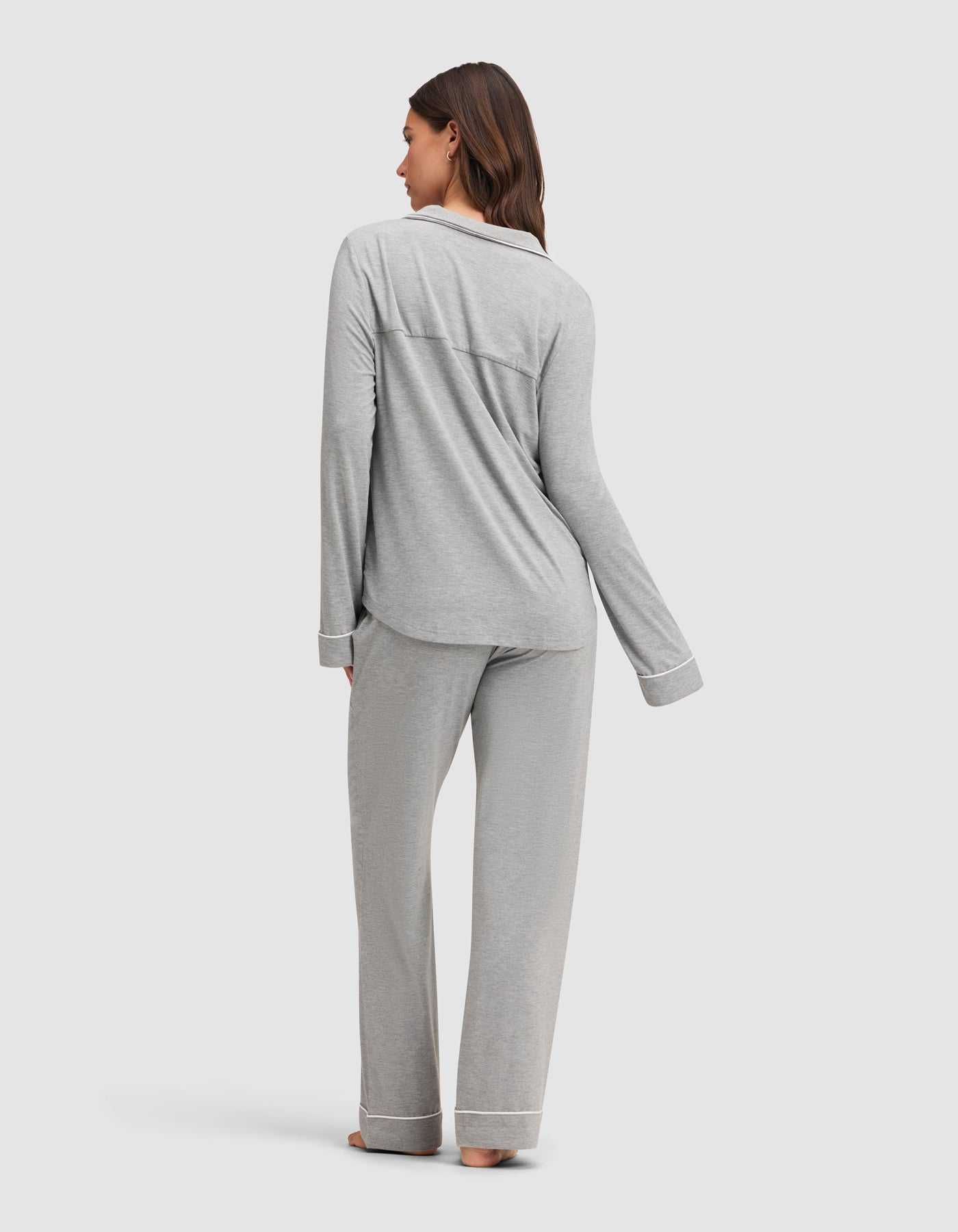A woman stands barefoot, facing away, wearing the Cozy Earth Women's Bamboo Stretch-Knit Long Sleeve Pajama Set in light gray with white trim. She is on a plain light background. |Color:Grey
