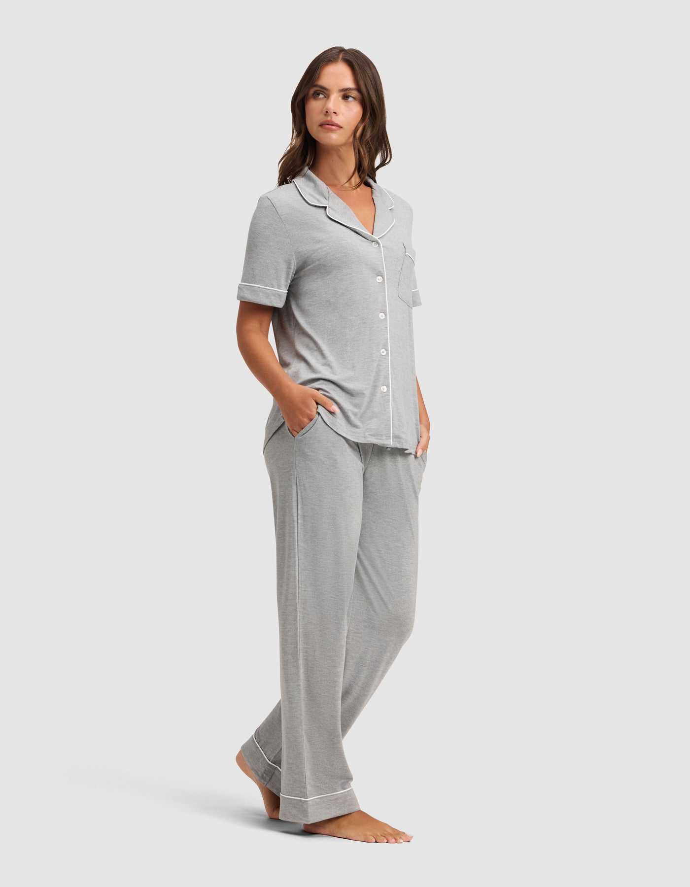 A woman stands barefoot in a Cozy Earth Women's Bamboo Stretch-Knit Short Sleeve & Pant Pajama Set, featuring light gray fabric with white piping and loose pants, hands in pockets, against a plain light background. |Color:Grey