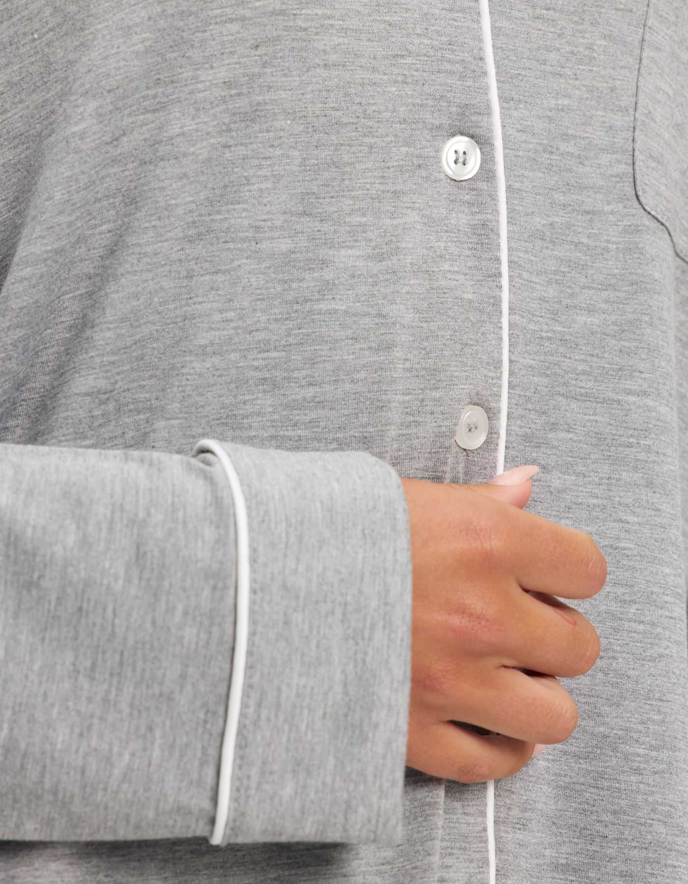 A close-up of a person’s hand holding the front of Cozy Earth’s Women’s Bamboo Stretch-Knit Long Sleeve Pajama Set in light grey, featuring white piping and buttons. The soft bamboo fabric looks comfortable and cozy. |Color:Grey