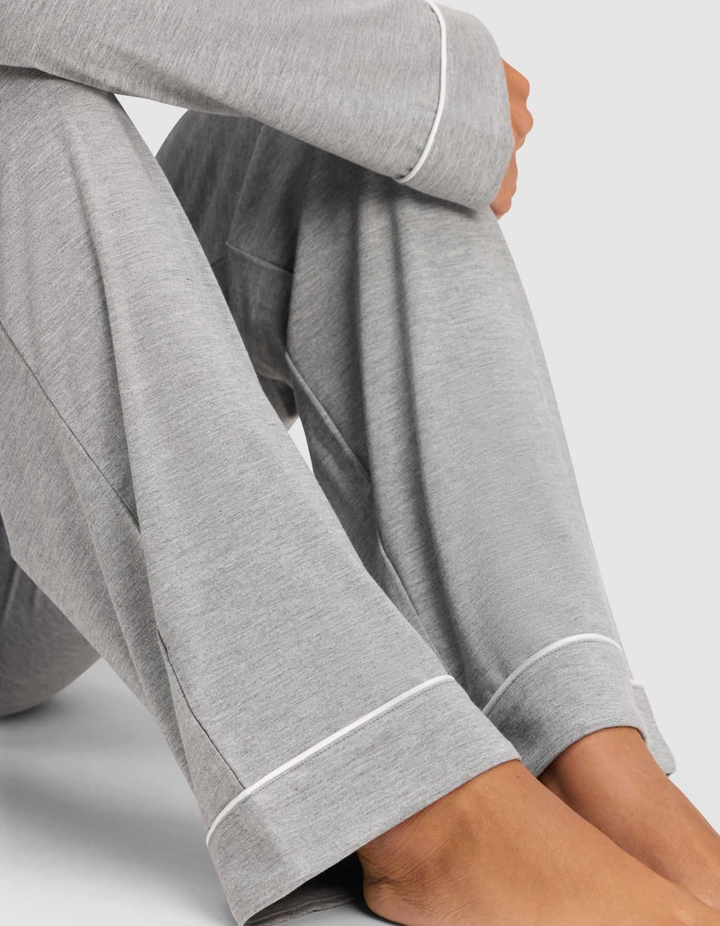 A close-up shows a person sitting cross-legged in the Cozy Earth Women's Bamboo Stretch-Knit Long Sleeve Pajama Set, light gray with white piping at the cuffs, barefoot against a plain background. |Color:Grey