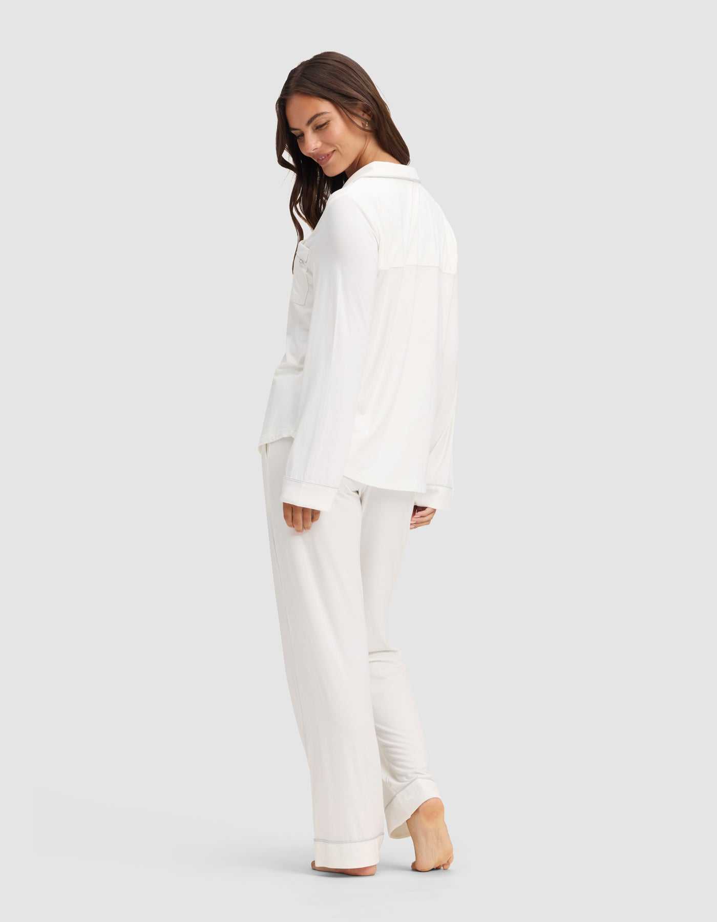 A woman with long brown hair stands barefoot, smiling and turning slightly to the side. She wears the Cozy Earth Women's Bamboo Stretch-Knit Long Sleeve Pajama Set in white, with pants, against a plain light gray background. |Color:Ivory