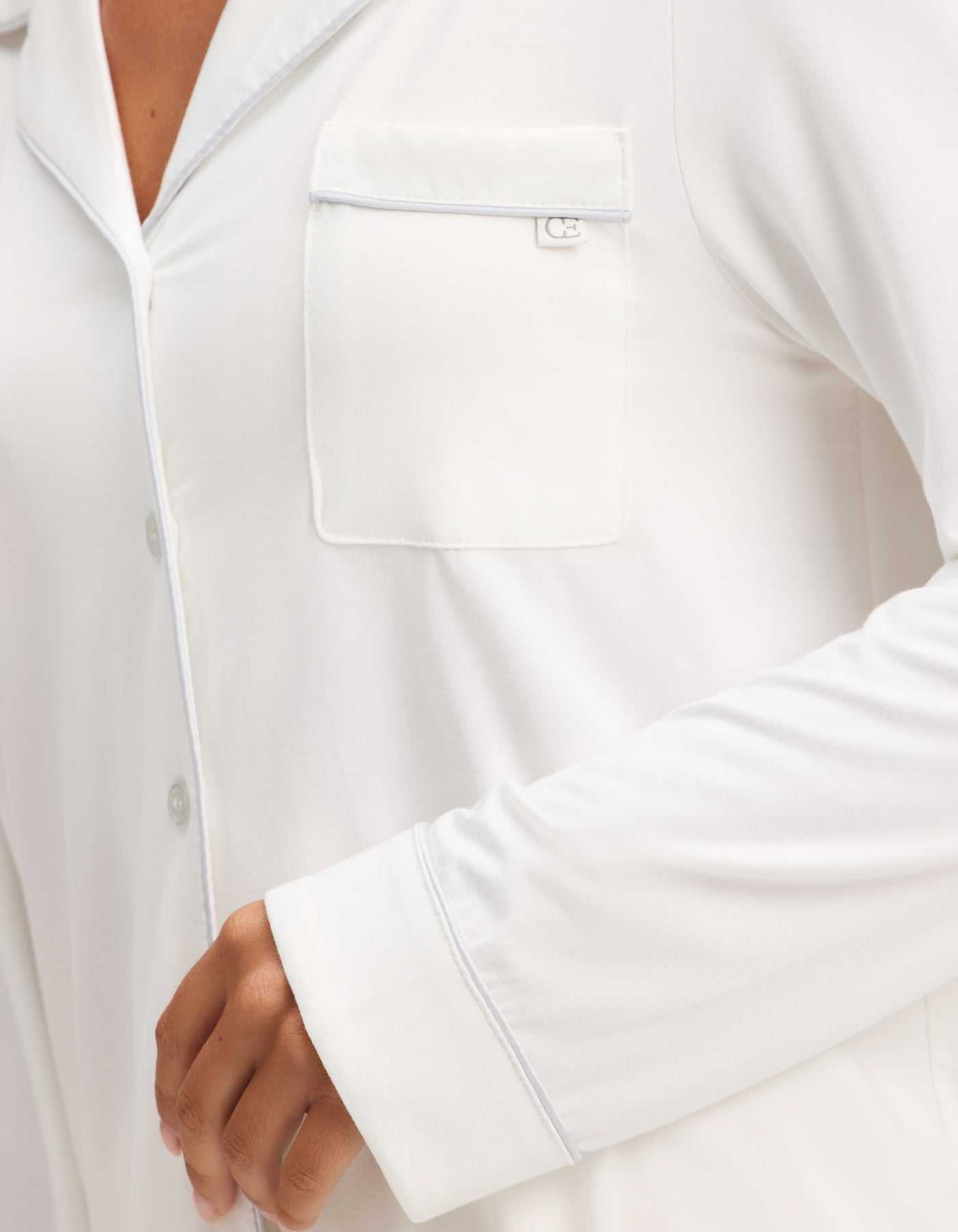 A close-up shows a person wearing the Cozy Earth Women's Bamboo Stretch-Knit Long Sleeve Pajama Set, featuring a white button-up top with a chest pocket and light piping. Only part of the torso and bent arm are visible. |Color:Ivory