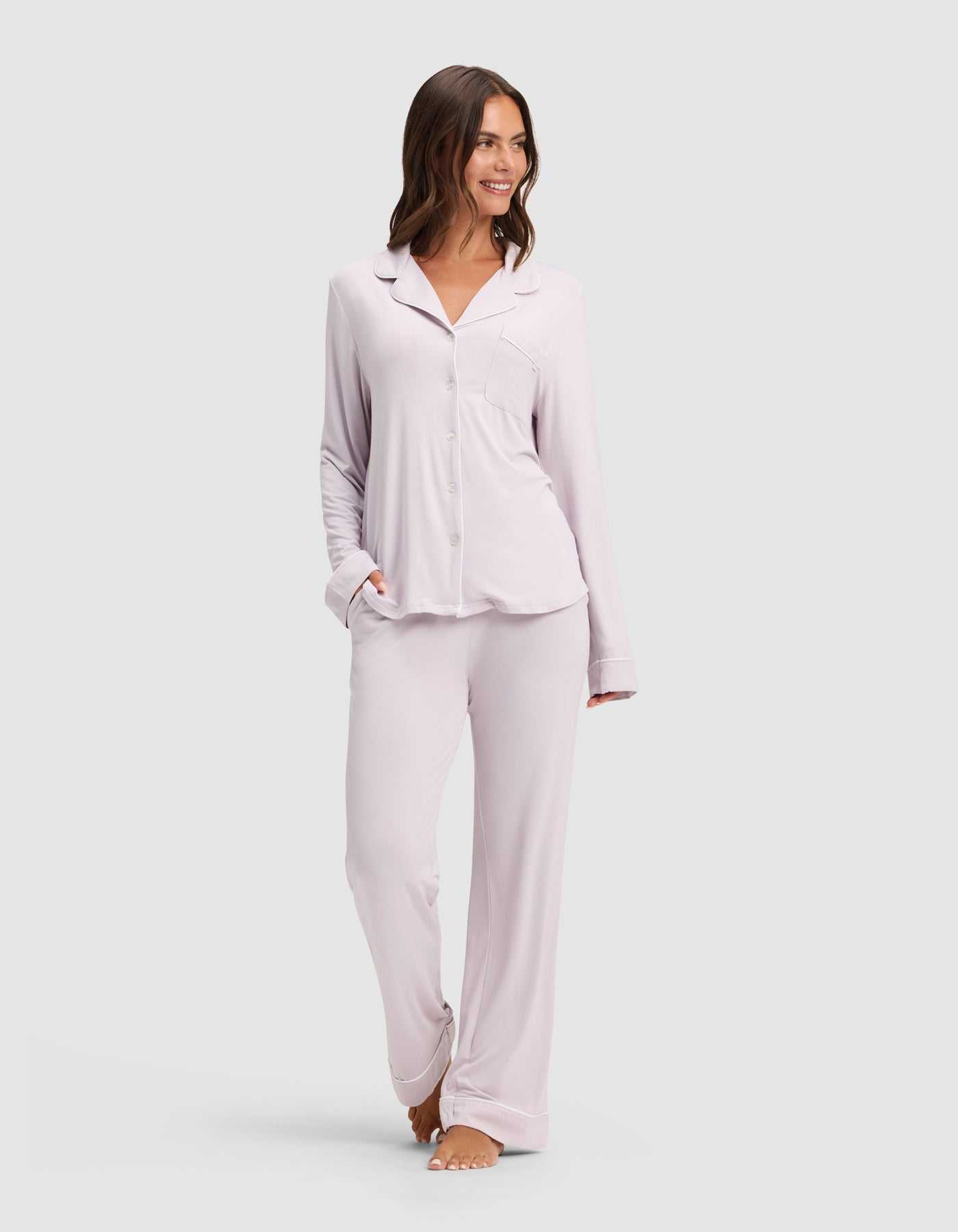 A woman smiles while wearing the Cozy Earth Women's Bamboo Stretch-Knit Long Sleeve Pajama Set, featuring a button-up top and matching pants in light lavender, standing barefoot against a plain light background. |Color:Lilac