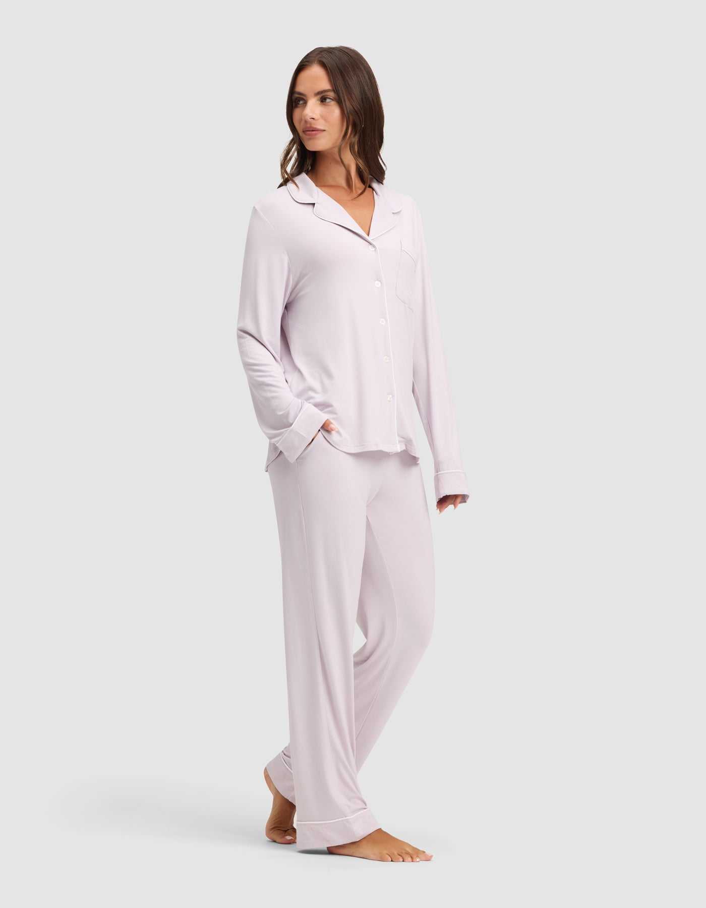 A woman wears the Cozy Earth Women's Bamboo Stretch-Knit Long Sleeve Pajama Set in light purple with matching pants, standing barefoot on a light gray background and looking to the side with one hand in her pocket. |Color:Lilac