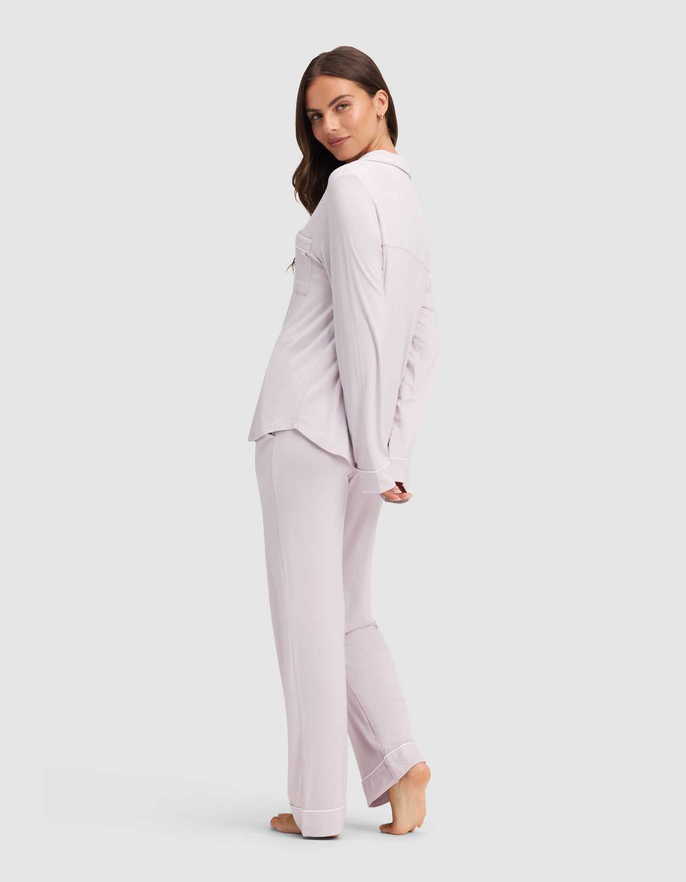 A woman stands barefoot on a light background, turned slightly to the side and looking over her shoulder. She wears the Cozy Earth Women's Bamboo Stretch-Knit Long Sleeve Pajama Set in matching light lavender. |Color:Lilac