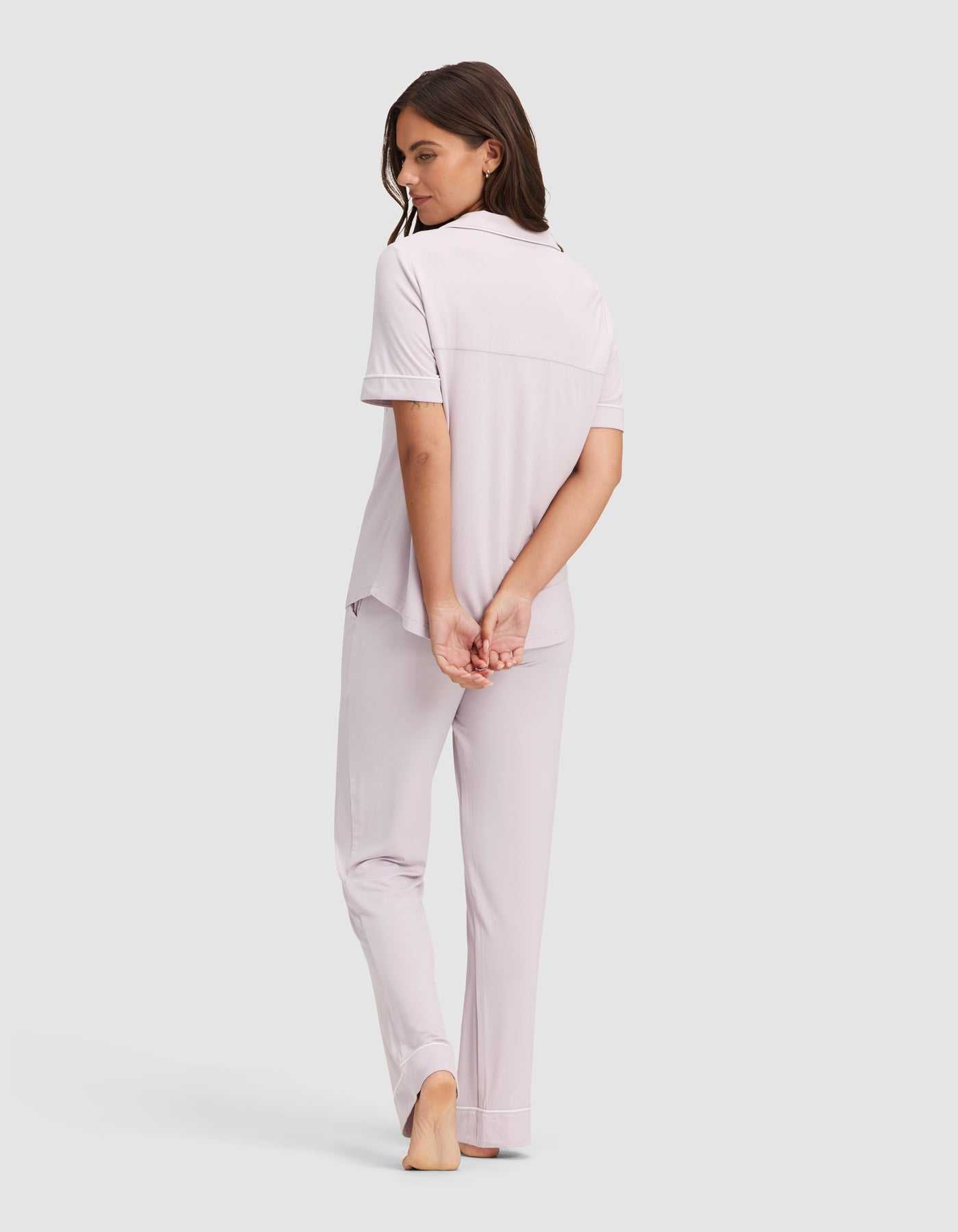 A woman models the Cozy Earth Women's Bamboo Stretch-Knit Short Sleeve & Pant Pajama Set in light lavender, standing barefoot with her hands behind her back, looking over her shoulder against a plain light background.|Color:Lilac