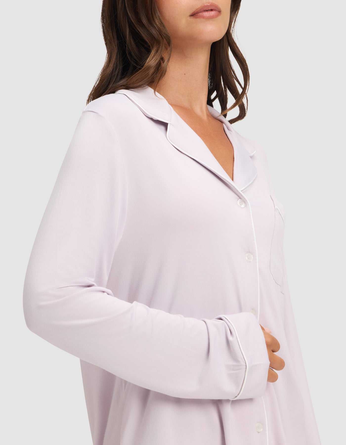 A woman wears the Cozy Earth Women's Bamboo Stretch-Knit Long Sleeve Pajama Set in light pink with white piping, standing against a plain background with one hand on her abdomen. Her face is partially out of frame. |Color:Lilac