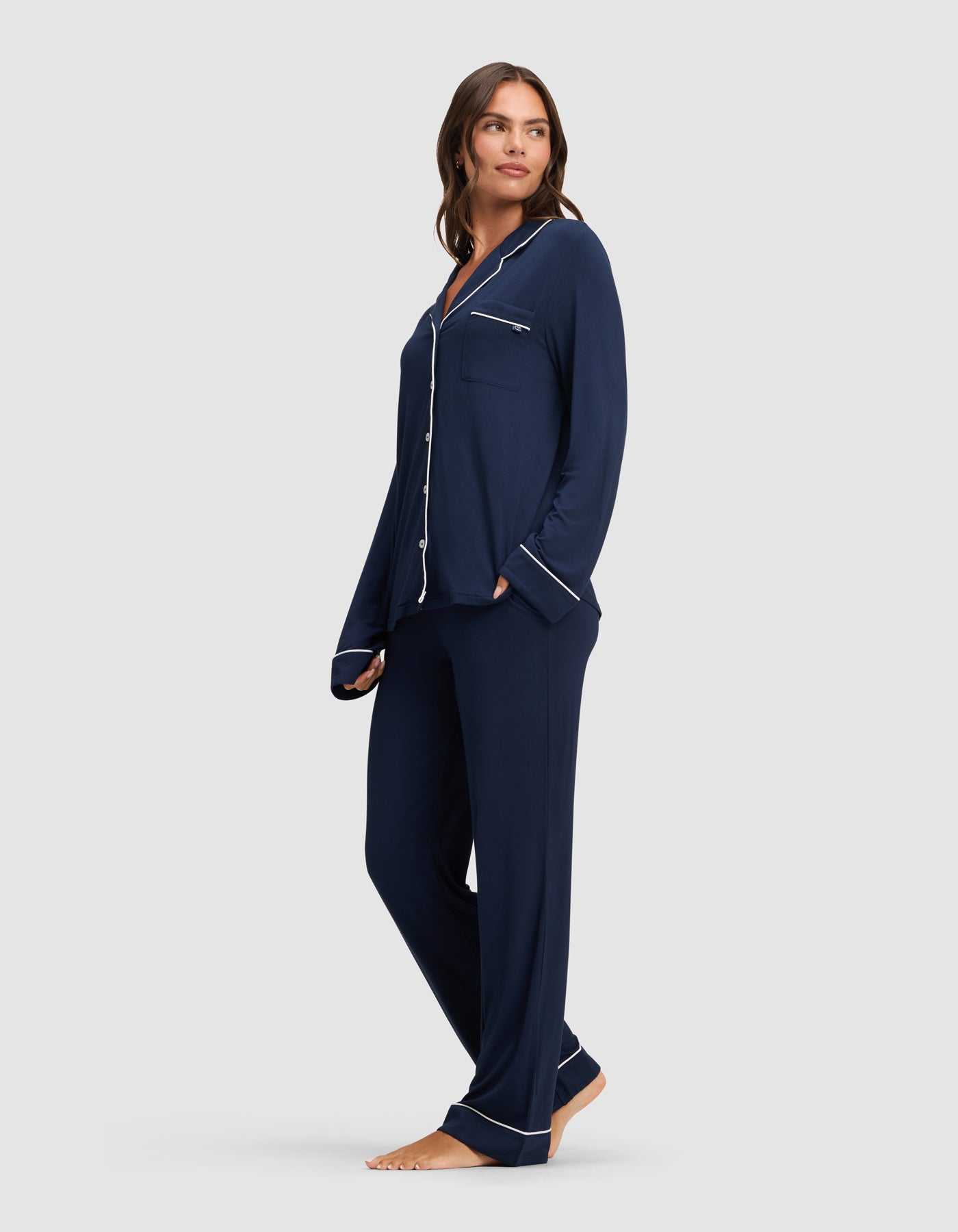 A woman stands barefoot against a plain light gray background, wearing the Cozy Earth Women's Bamboo Stretch-Knit Long Sleeve Pajama Set in navy with white piping, featuring a button-up top and matching pants.|Color:Navy