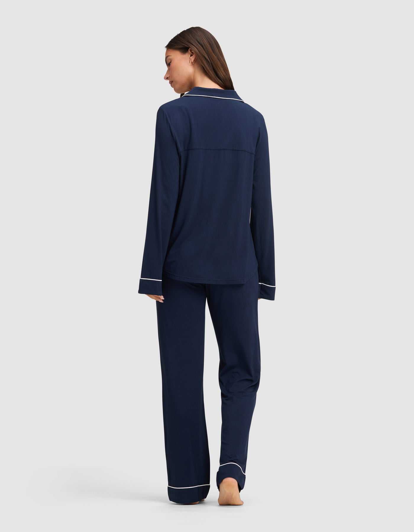 A woman, barefoot and facing away, wears the Cozy Earth Women's Bamboo Stretch-Knit Long Sleeve Pajama Set in navy blue with white piping on the cuffs and hem, set against a plain light gray background.|Color:Navy
