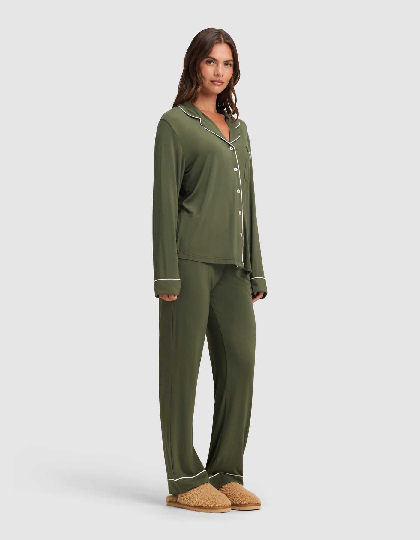A woman wears the Cozy Earth Women's Bamboo Stretch-Knit Long Sleeve Pajama Set in olive green with matching pants and tan slippers, standing against a plain light gray background. |Color:Olive