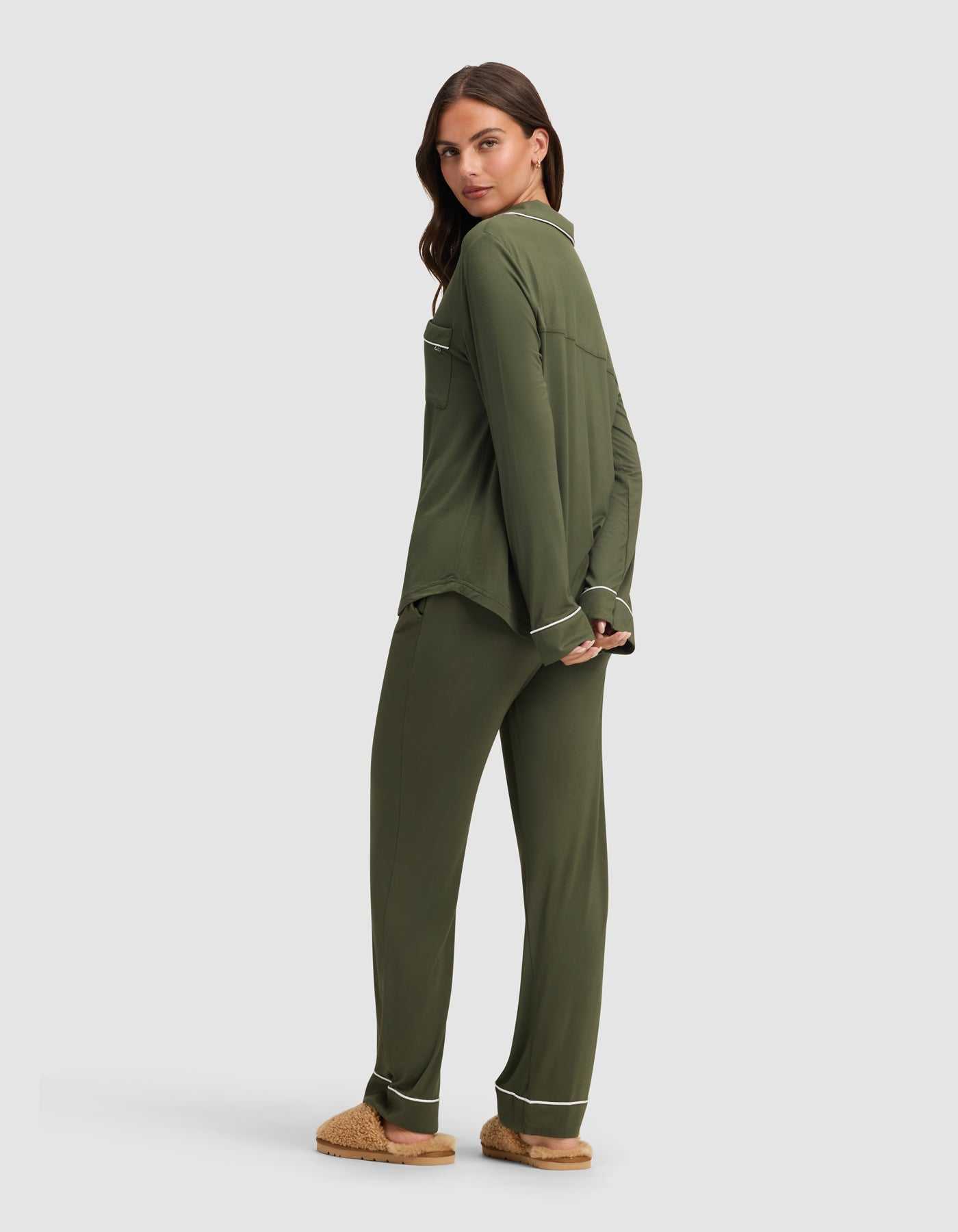 A woman in the Cozy Earth Women's Bamboo Stretch-Knit Long Sleeve Pajama Set in olive green with white piping and tan slippers stands against a light gray background, facing backward and looking over her shoulder. |Color:Olive