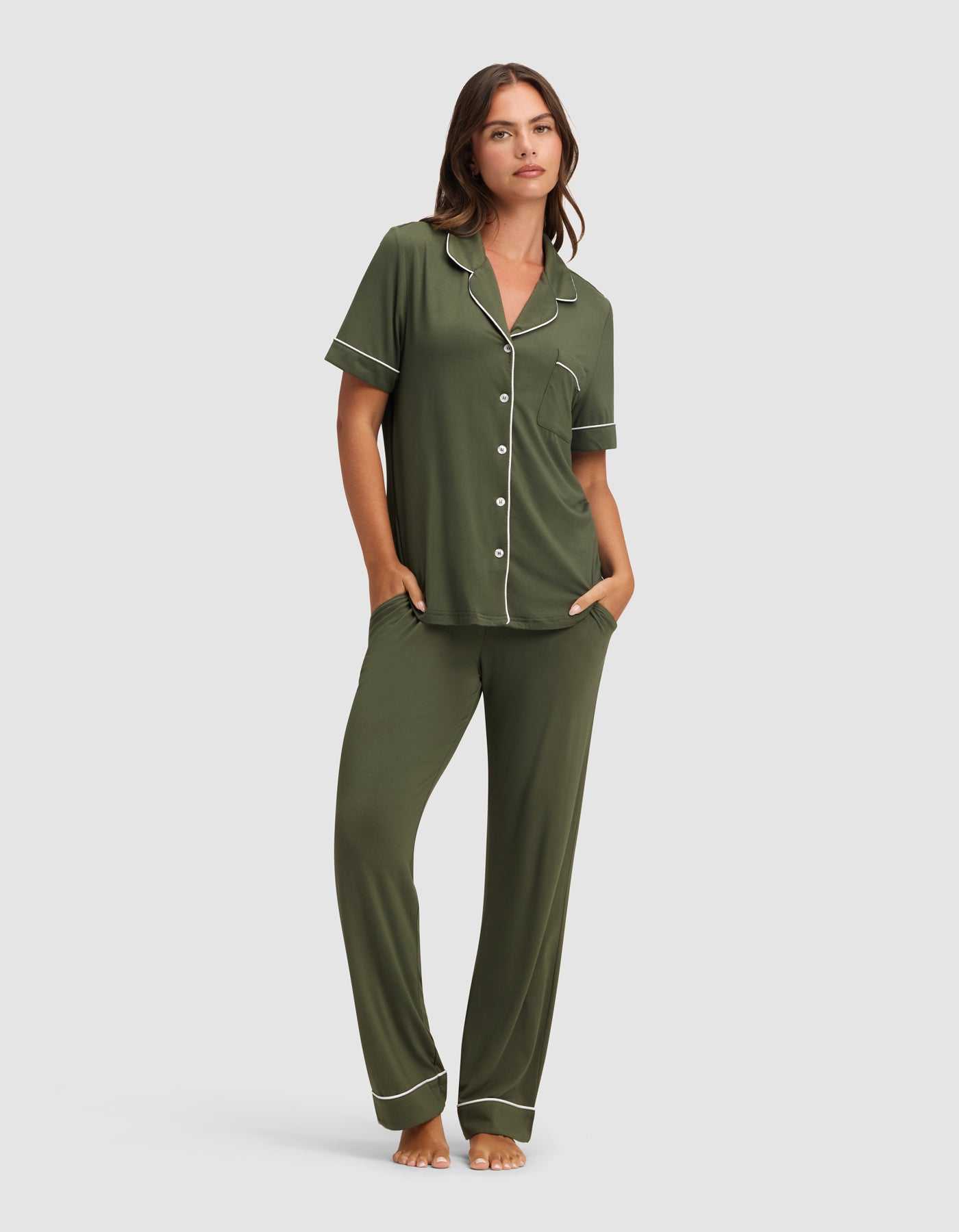 A woman stands barefoot in the Cozy Earth Women's Bamboo Stretch-Knit Short Sleeve & Pant Pajama Set in olive green with white piping, featuring a button-up top and long pants, her hands in her pockets against a plain light background.|Color:Olive