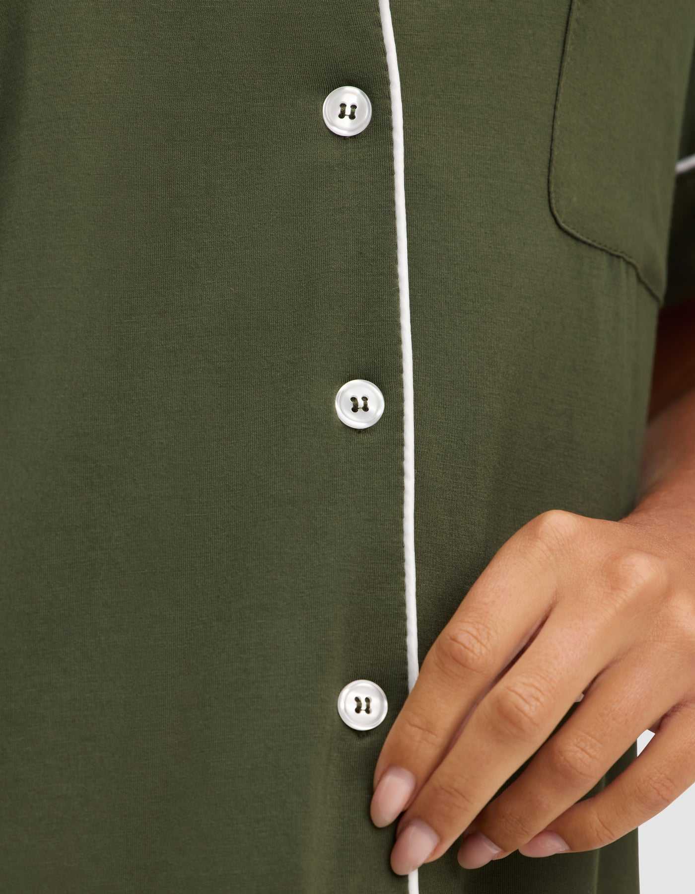 A hand gently touches the front of the HIDE Women's Bamboo Stretch-Knit Short Sleeve Pajama Top in olive green, featuring white buttons and white piping near the buttons and pocket. |Color:Olive