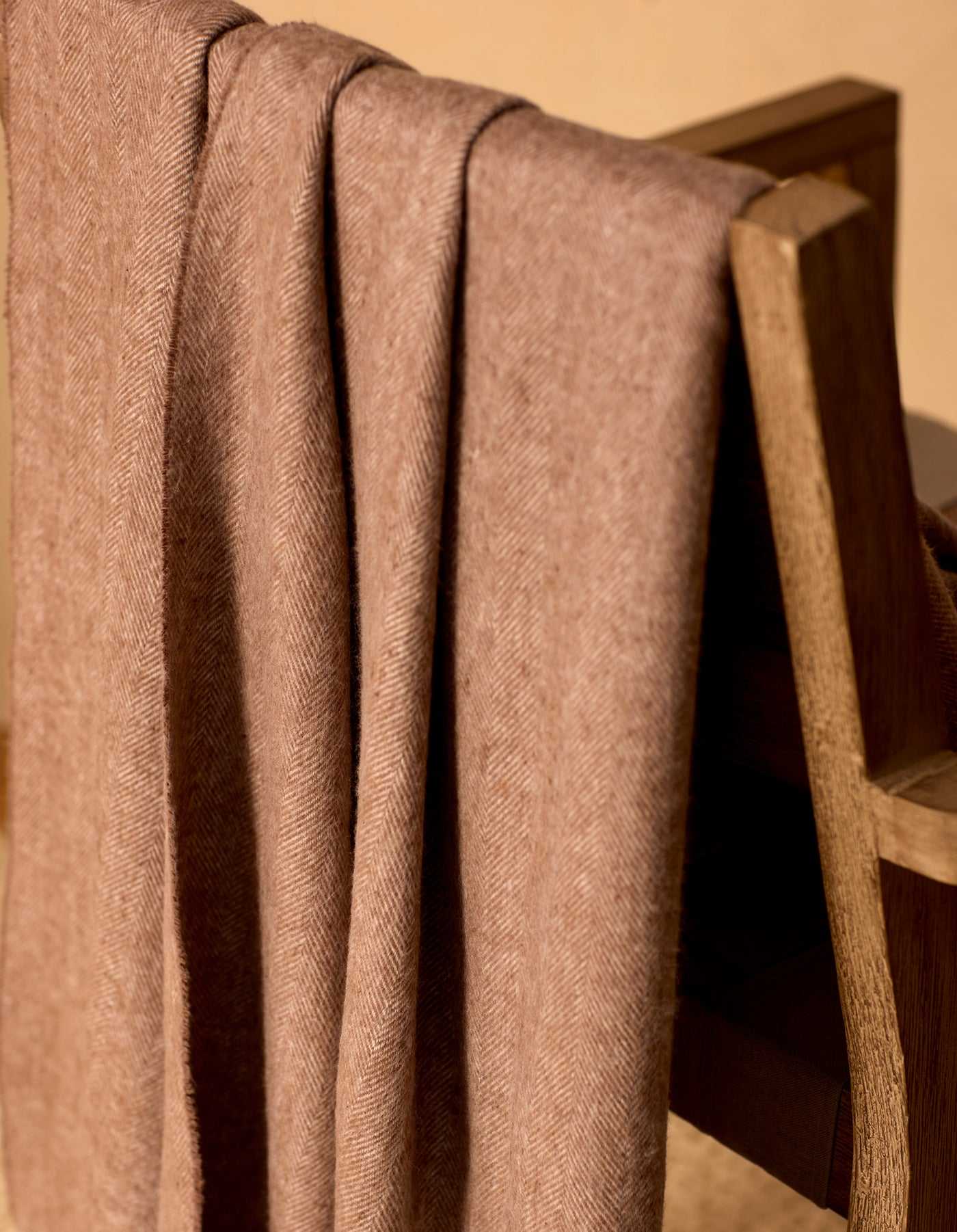 The Cozy Earth Bamboo Herringbone Tassel Throw, in light brown with a herringbone pattern, is draped neatly over a wooden chair, bathed in warm lighting.|Color:Truffle