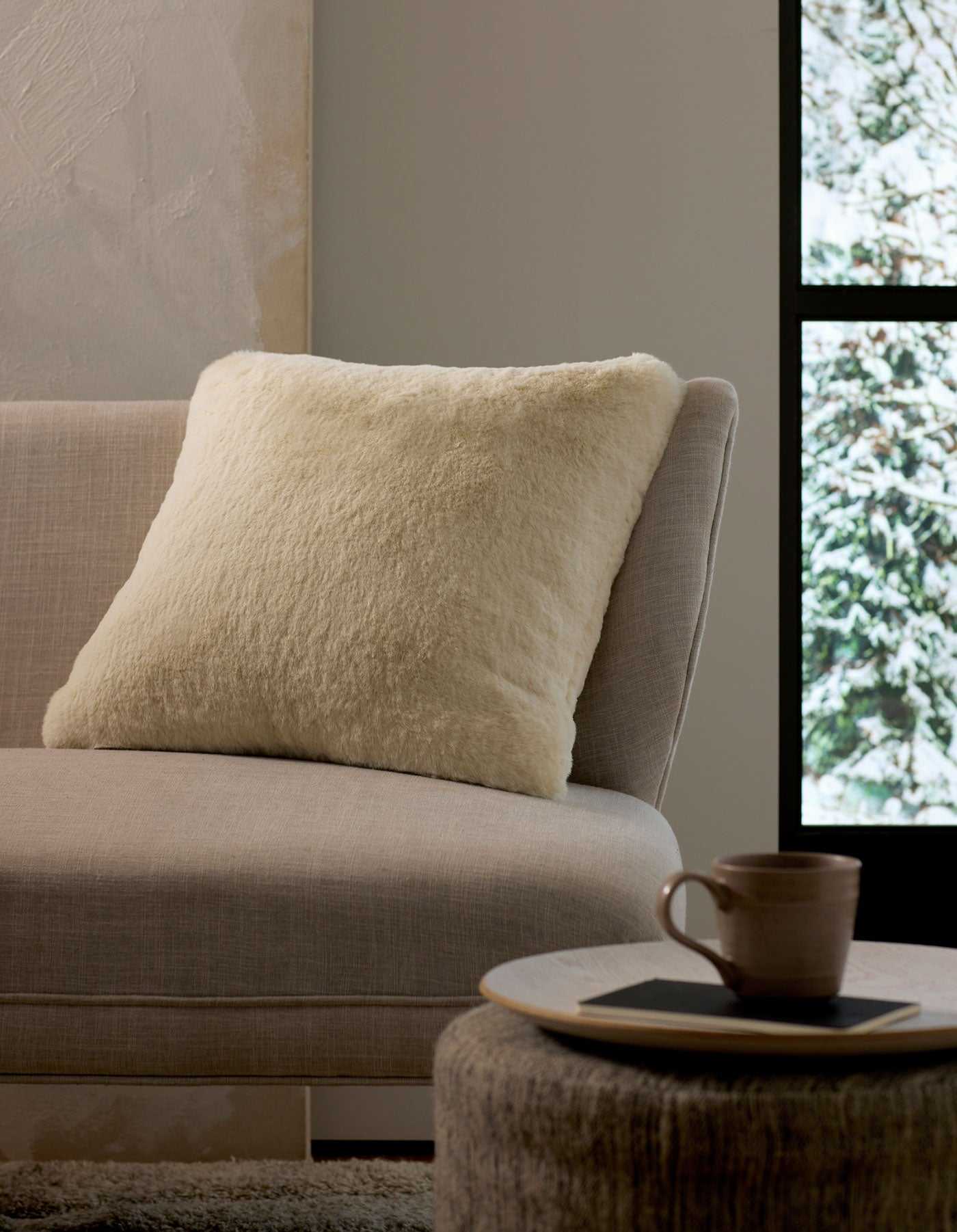 A cozy interior scene with a light beige sofa, a fluffy Cozy Earth Cuddle Pillow in cream, a round ottoman holding a cup and saucer, and a window framing a snowy outdoor view.|Color:Creme