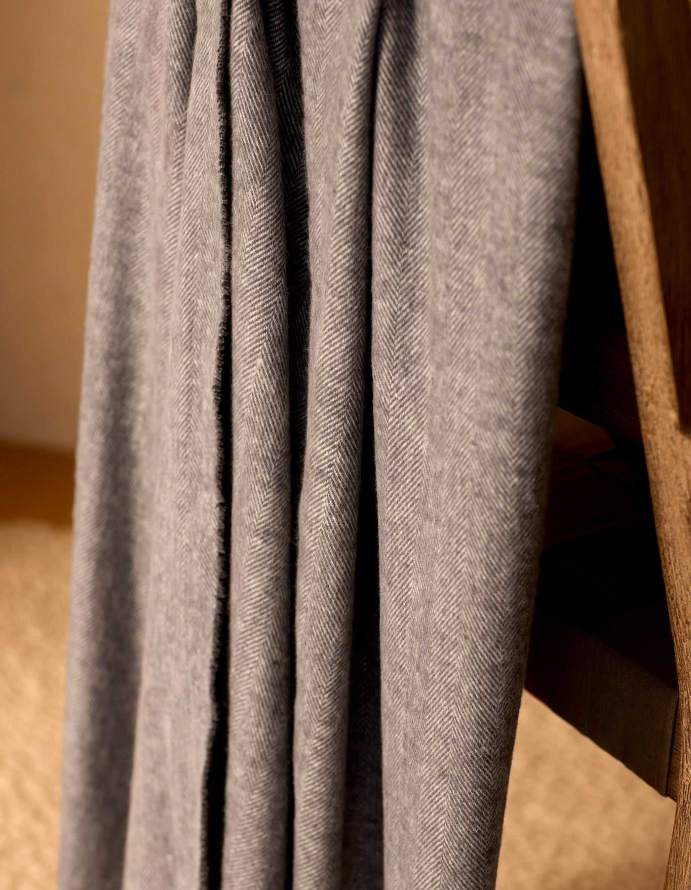A close-up of the Cozy Earth Bamboo Herringbone Tassel Throw, featuring a gray herringbone pattern, draped over a wooden chair against a blurred beige background.|Color:Shadow
