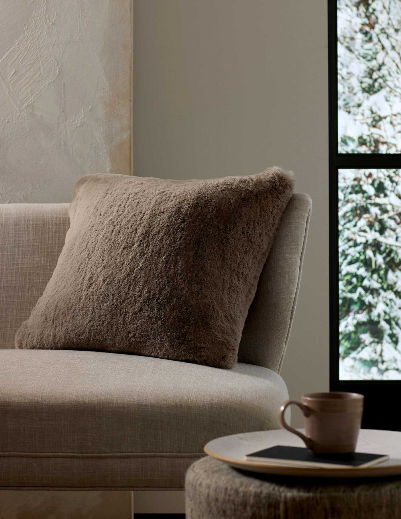 A cozy, neutral-toned living space features the Cozy Earth Cuddle Pillow in taupe on a beige sofa next to a round side table with a ceramic mug and book, framed by a window view of snowy trees.|Color:Walnut