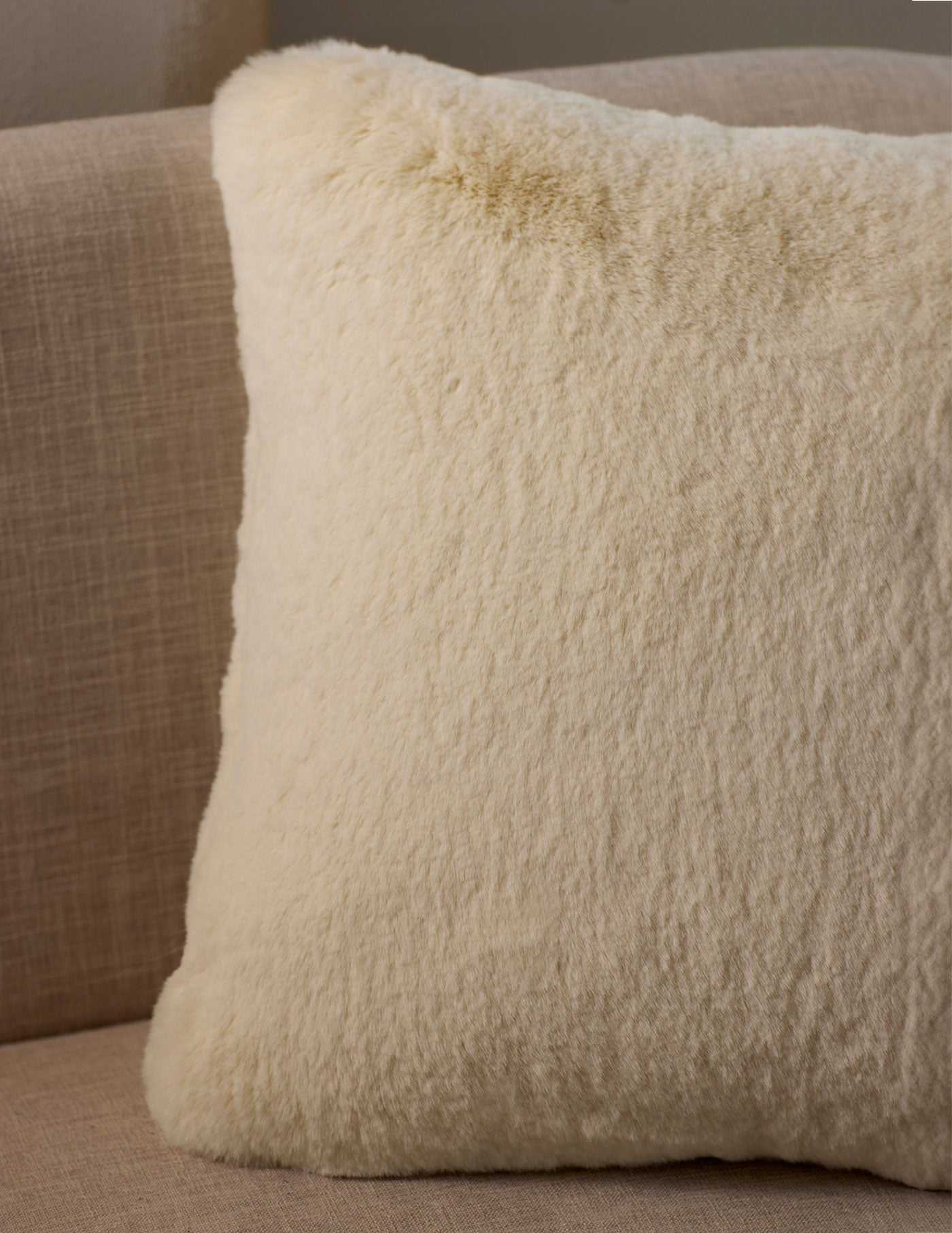 A close-up of the Cuddle Pillow by Cozy Earth, featuring a fluffy cream color and soft texture, placed on a light beige fabric sofa.|Color:Creme