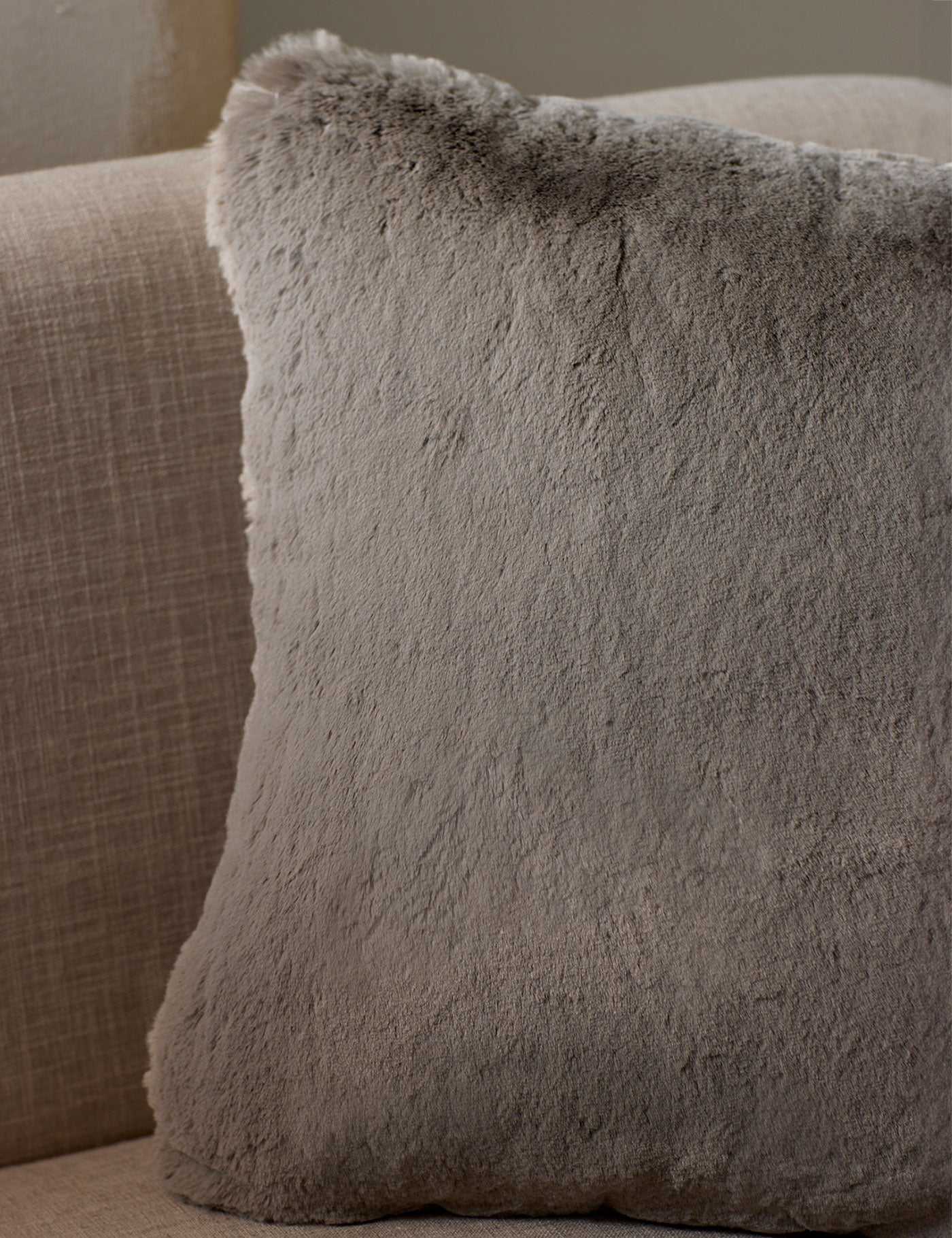 The Cozy Earth Cuddle Pillow, a large square in light gray faux fur, rests against the back of a beige fabric sofa.|Color:Dove Grey