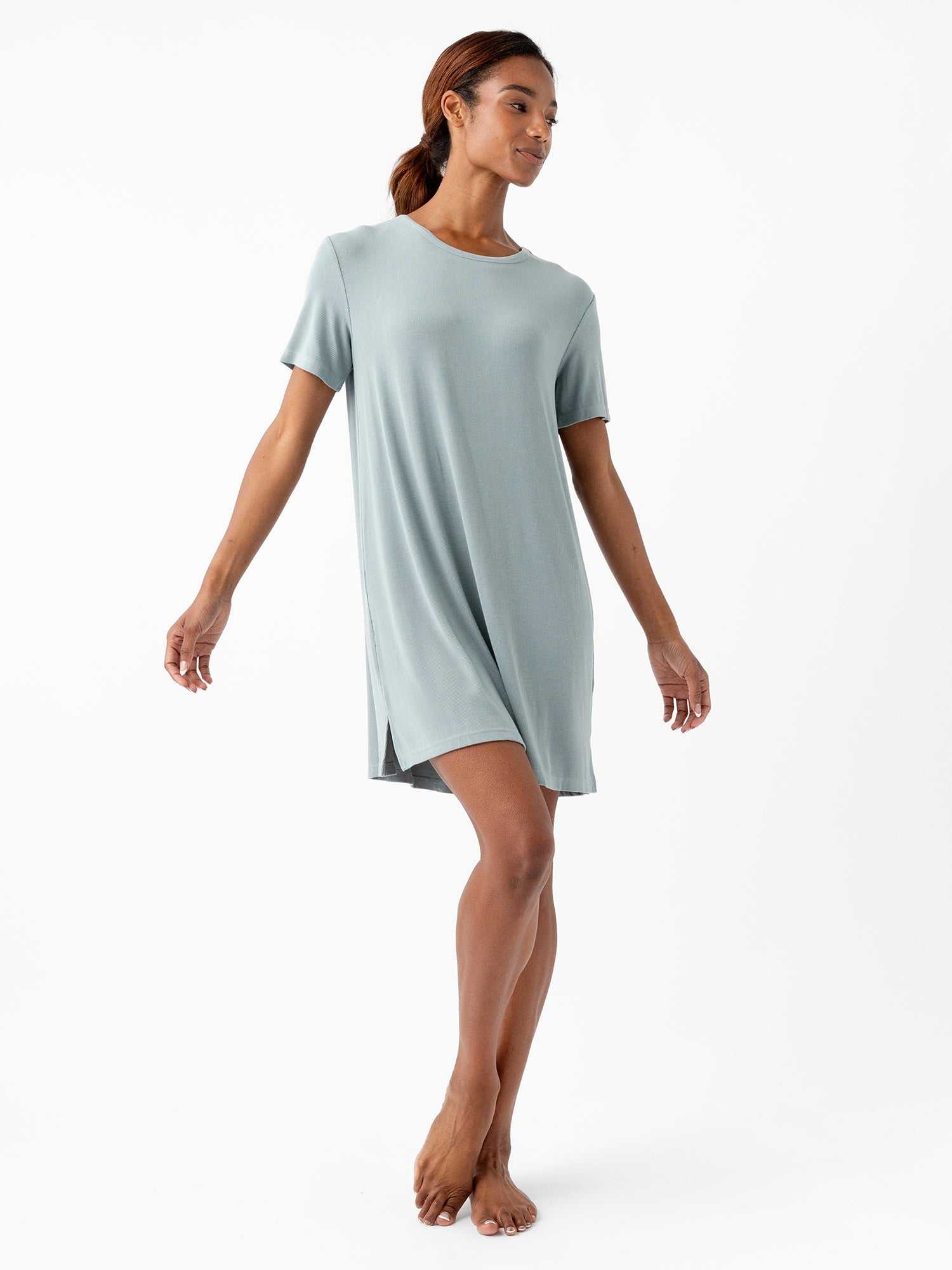 A woman stands barefoot on a white background, wearing the loose-fitting, short-sleeved Women’s Bamboo Rib-Knit Boyfriend Sleep Dress by Cozy Earth. Her head is slightly turned to the side, and her arms are extended, giving a sense of balance and grace. The teal sleep dress features a small slit on one side. |Color:Smokey Blue