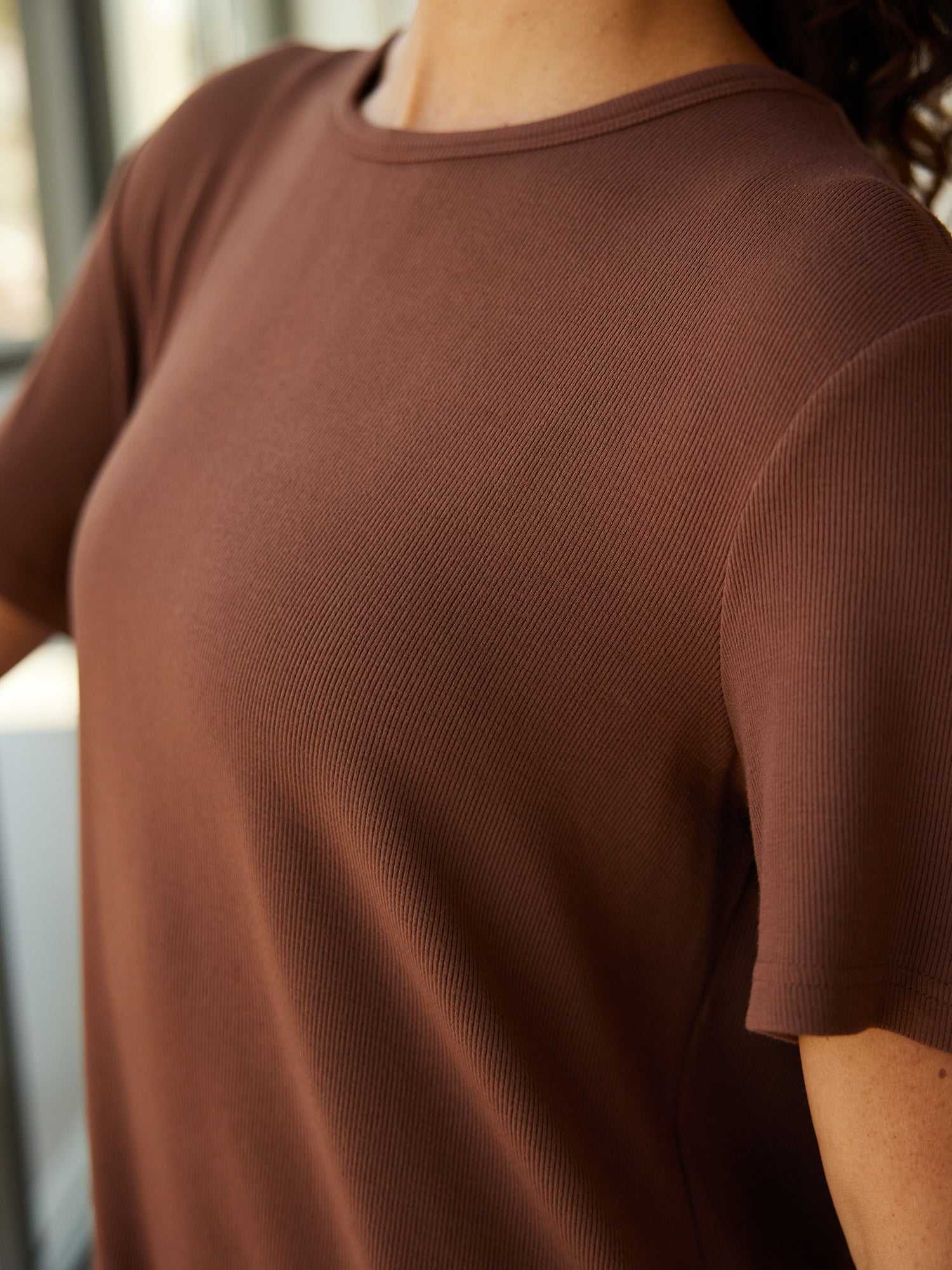 Close-up image of a person wearing Cozy Earth's Women’s Bamboo Rib-Knit Boyfriend Sleep Dress in brown. The focus is on the upper part of the sleep dress and the right shoulder, with the person's head not visible. The background appears to be indoors with blurred windows. |Color:Spice