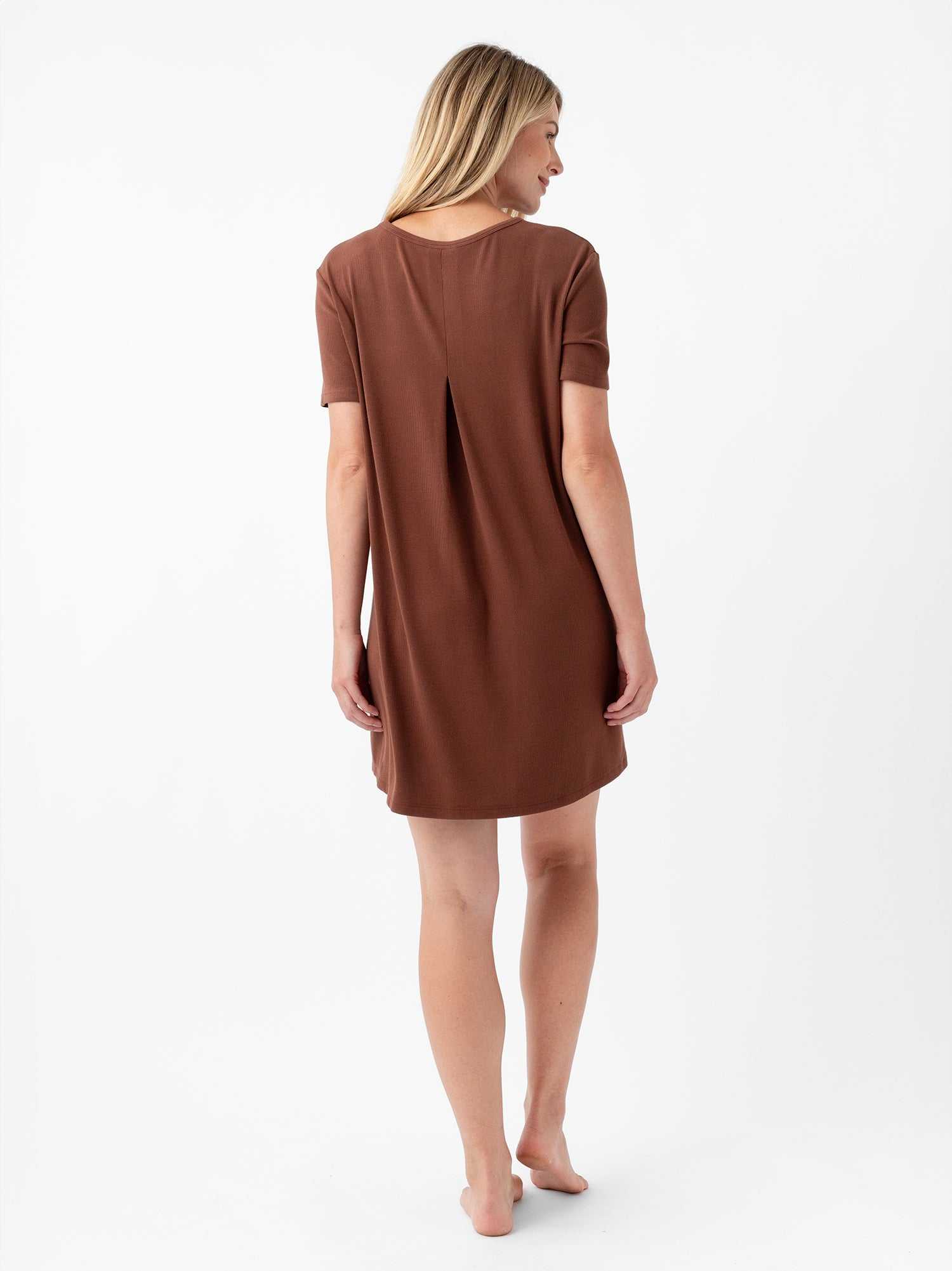 A woman with long blonde hair, standing barefoot, is shown from the back wearing the Cozy Earth Women’s Bamboo Rib-Knit Boyfriend Sleep Dress in a knee-length brown design with short sleeves and a central pleat detail. The plain white background highlights the simplicity and elegance of this dress. |Color:Spice