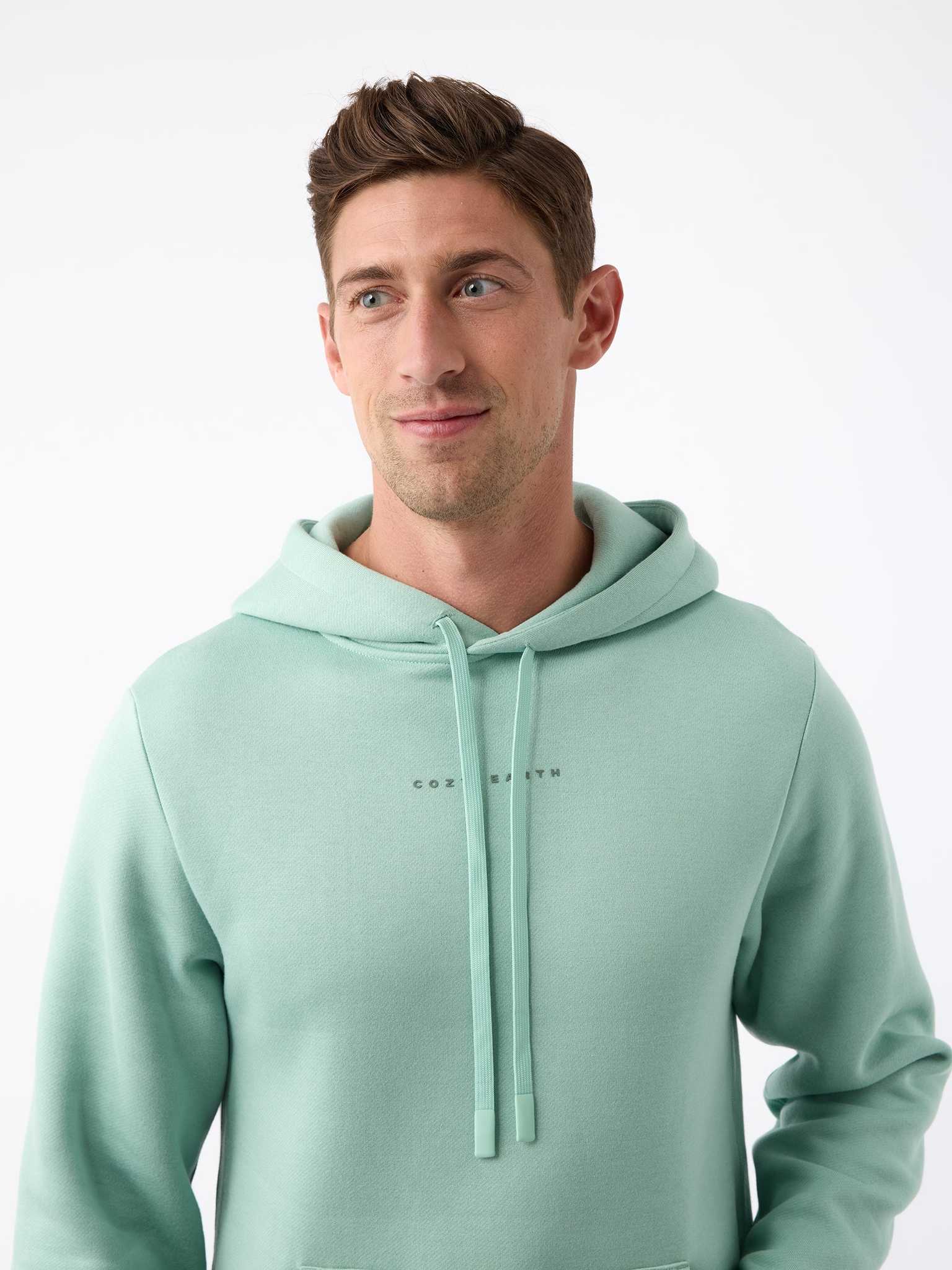 A person wearing the Men's CityScape Hoodie by Cozy Earth stands against a plain white background. The light green hoodie features drawstrings and subtle text on the chest. The person, with short brown hair, is looking slightly to the side while smiling gently. |Color:Wintergreen
