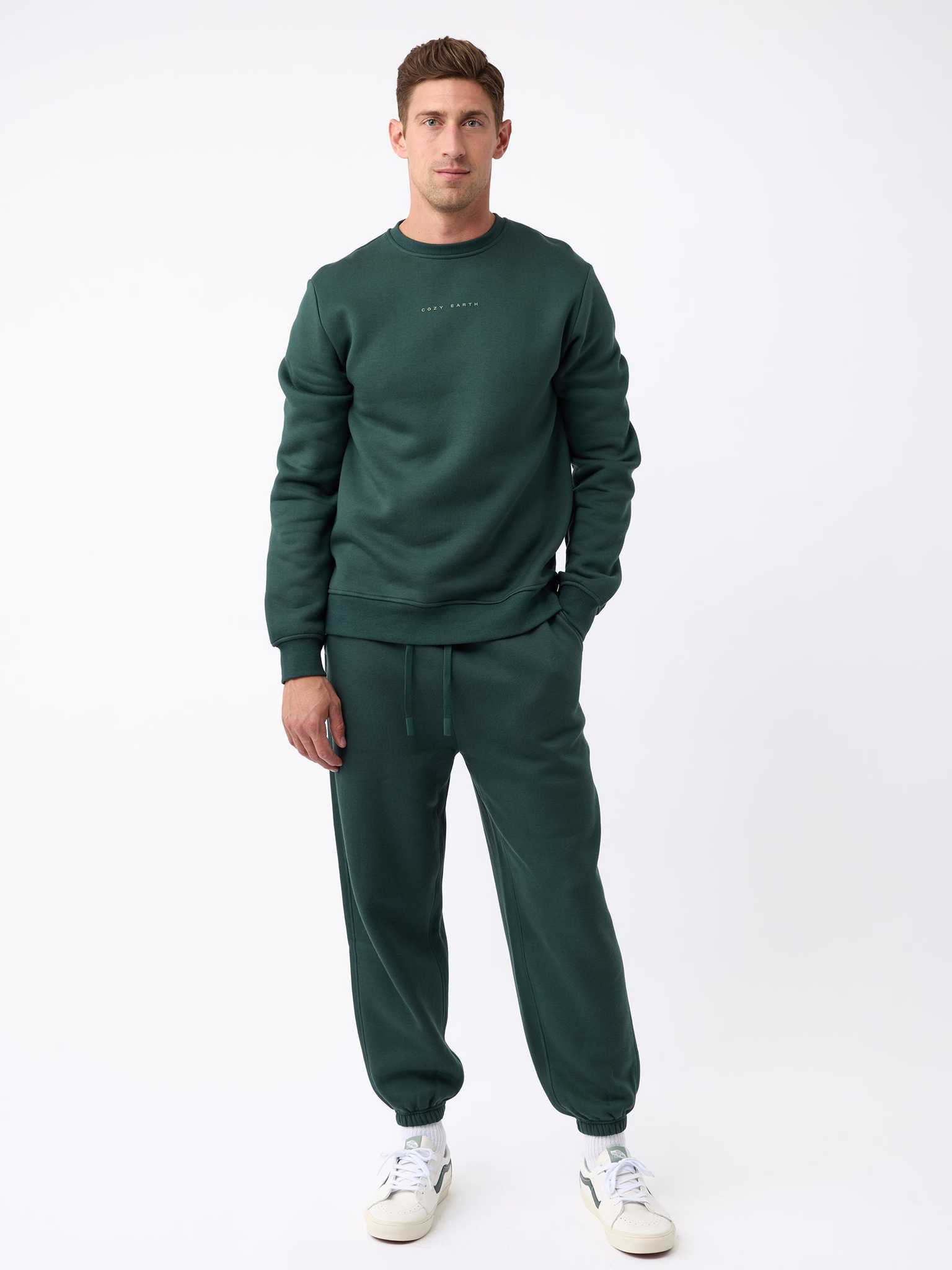 A man is standing against a white background wearing the dark green Men's CityScape Crewneck from Cozy Earth paired with matching joggers. He completes his look with white sneakers. |Color:Spruce