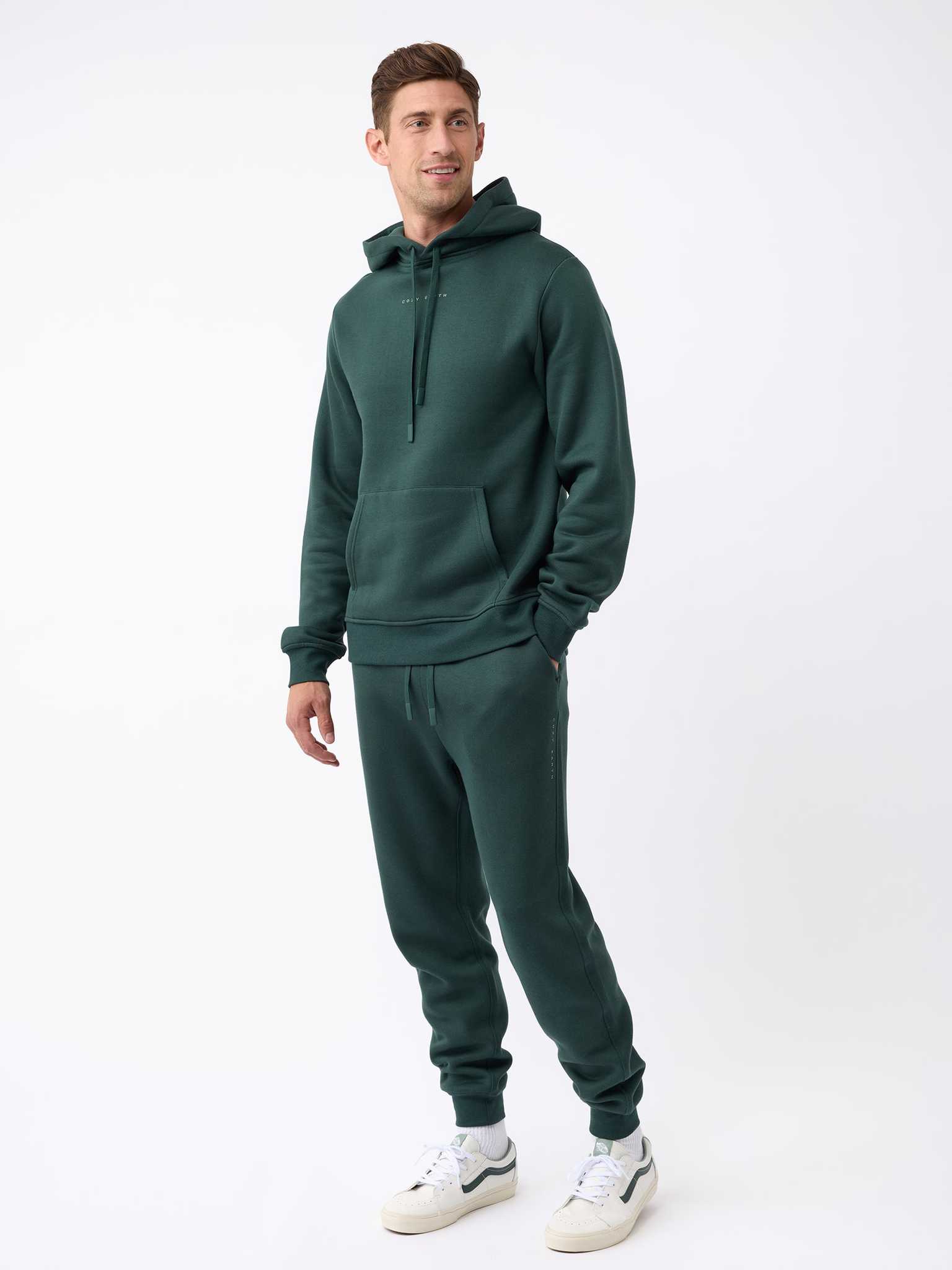 A person wearing the Men's CityScape Jogger by Cozy Earth in dark green stands against a plain white background. They are wearing white sneakers and have their hands in the jogger's hoodie pocket, looking slightly to the side. |Color:Spruce