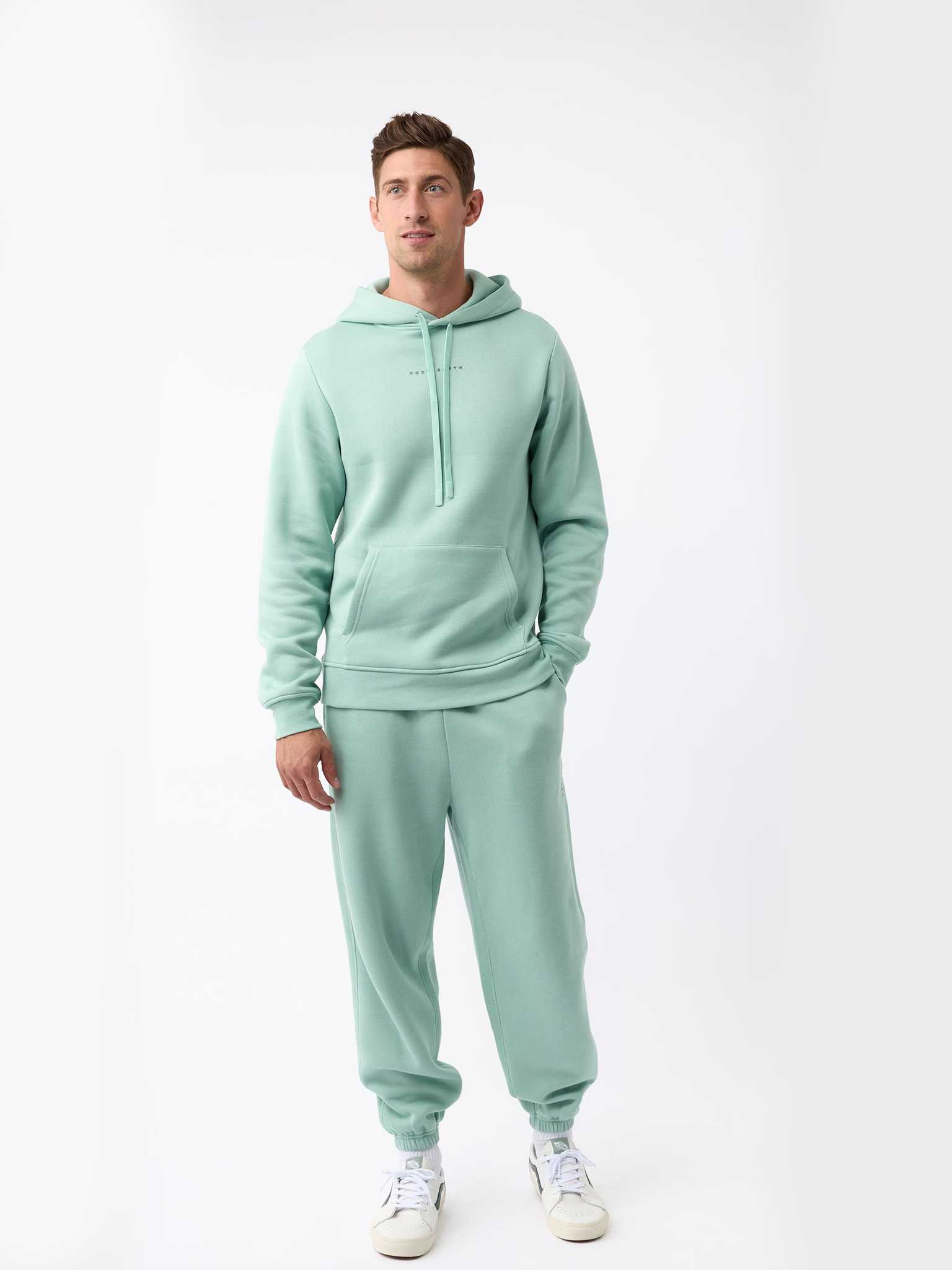A person is wearing the Men's CityScape Hoodie from Cozy Earth in mint green, paired with matching sweatpants, standing against a plain white background. They complete their outfit with white sneakers featuring green details. |Color:Wintergreen