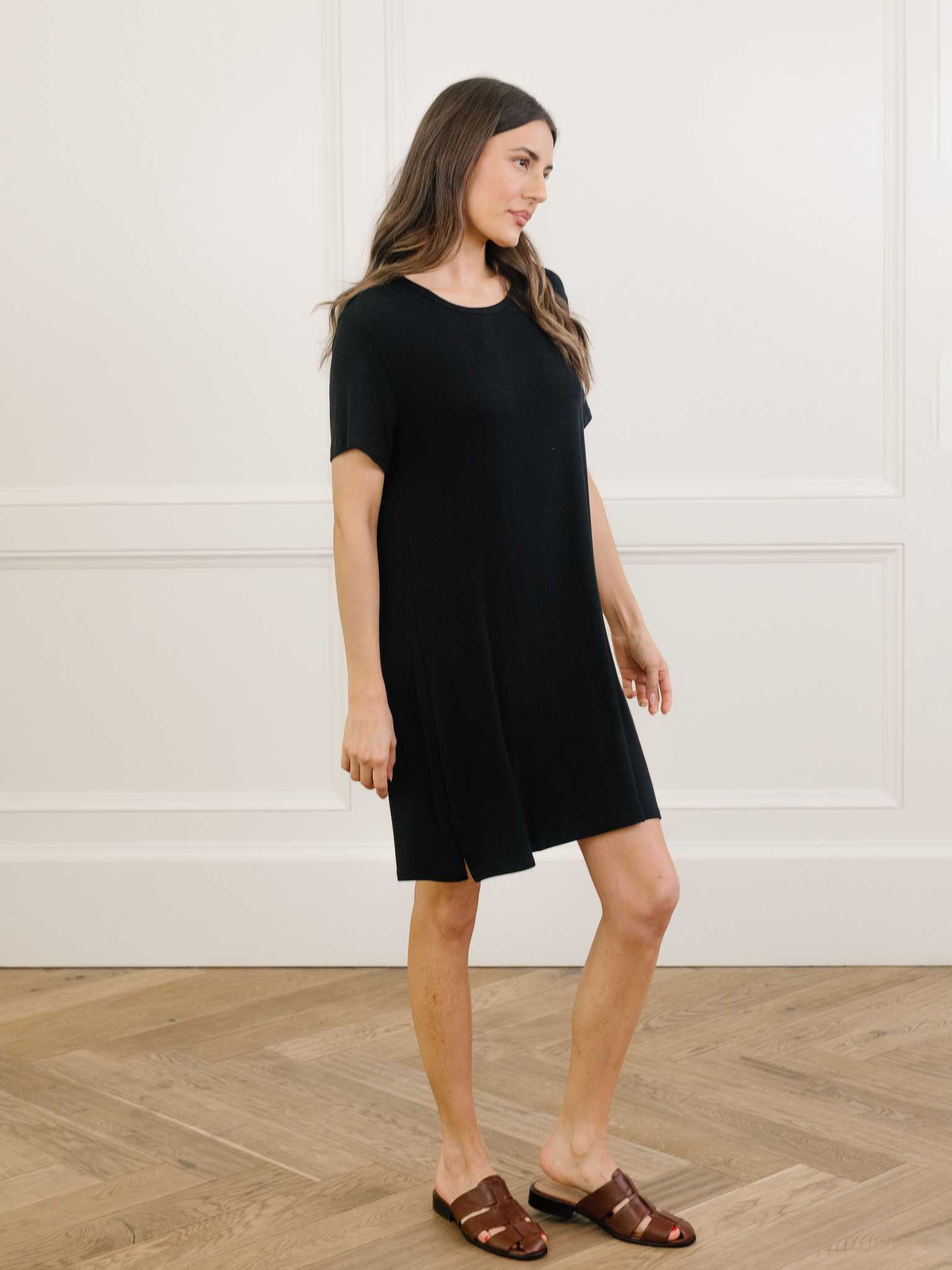 A woman standing in a black Cozy Earth bamboo rib-knit boyfriend sleep dress with short sleeves, knee-length, in a lounge or bedroom setting. |Color:Black