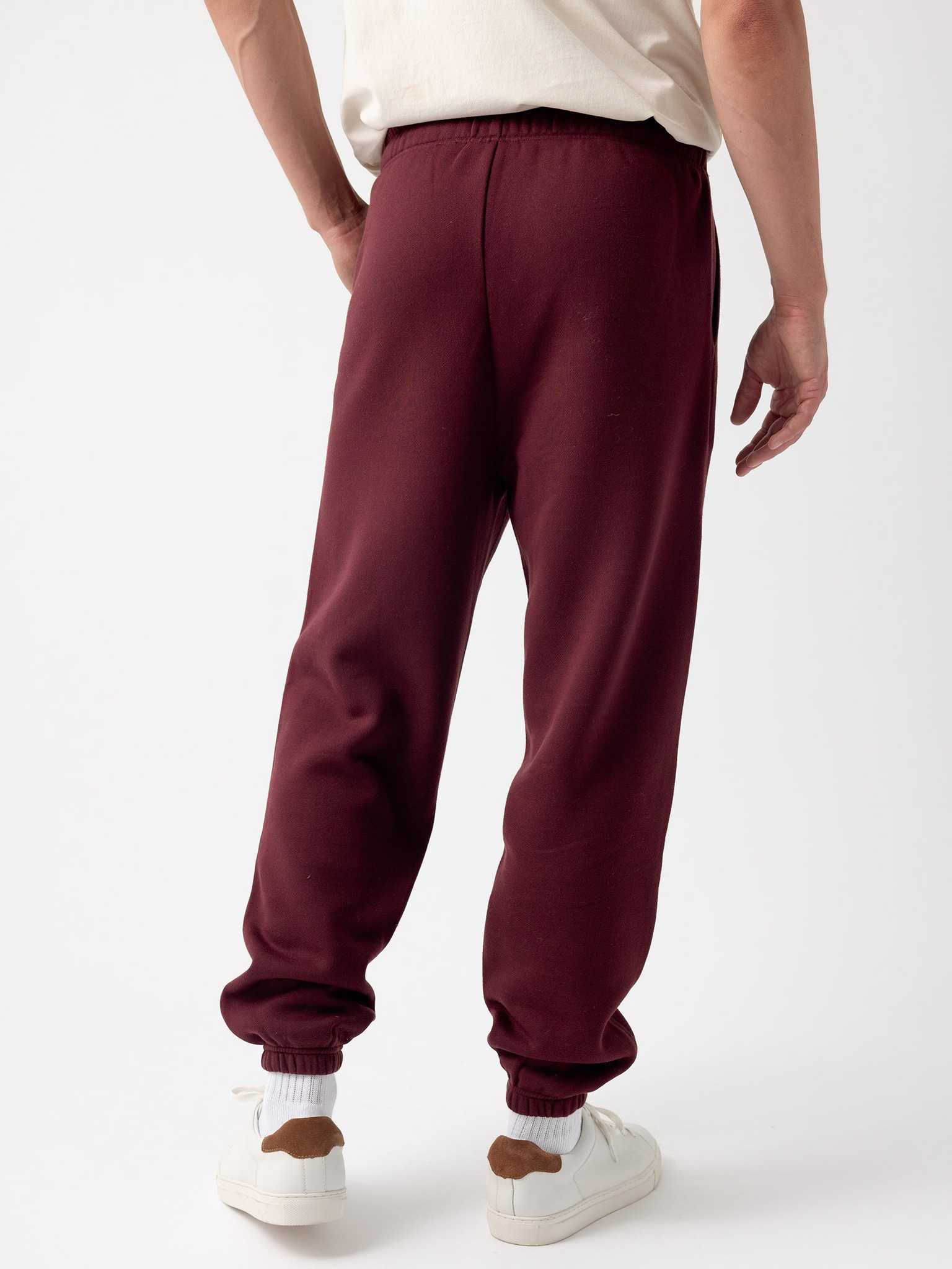 A person in a white shirt stands with their back to the camera, wearing the Cozy Earth Men's CityScape Sweatpant in maroon, which features elastic cuffs, paired with white sneakers that have brown accents. The background is plain white. |Color:Burgundy