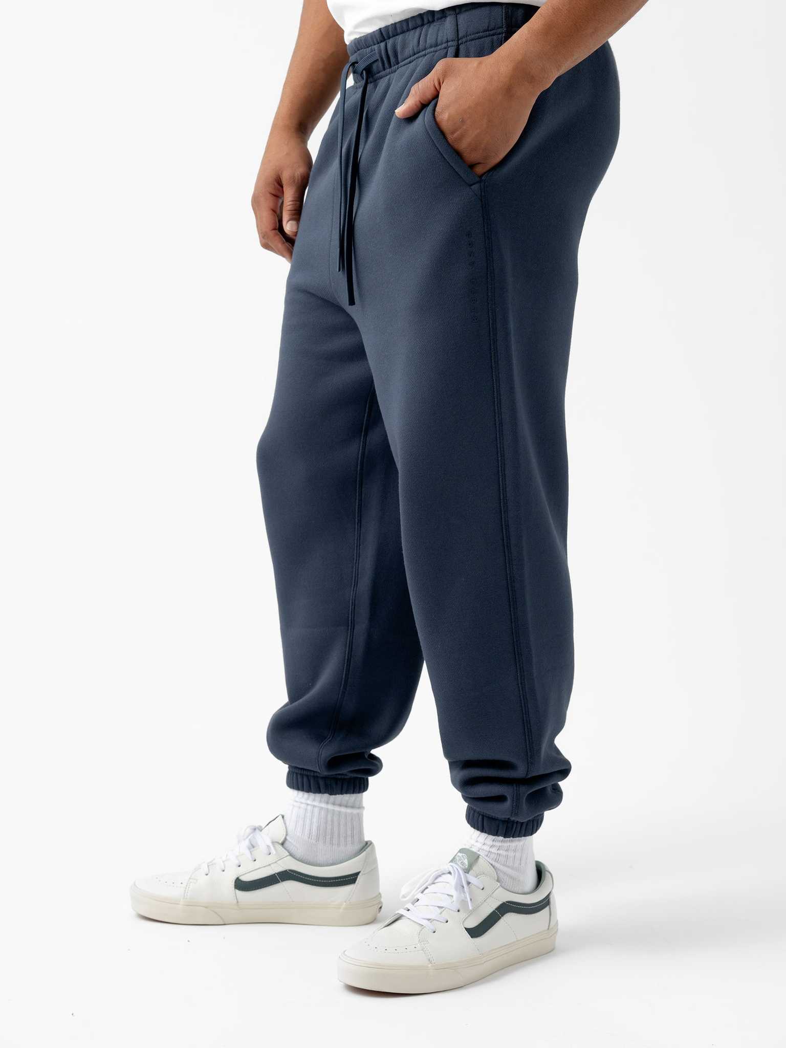 A person wearing Cozy Earth's Men's CityScape Sweatpant in dark blue with pockets, a white T-shirt, white socks, and white sneakers with dark accents stands against a plain white background. The person's hands are in the pockets of the sweatpants. |Color:Eclipse