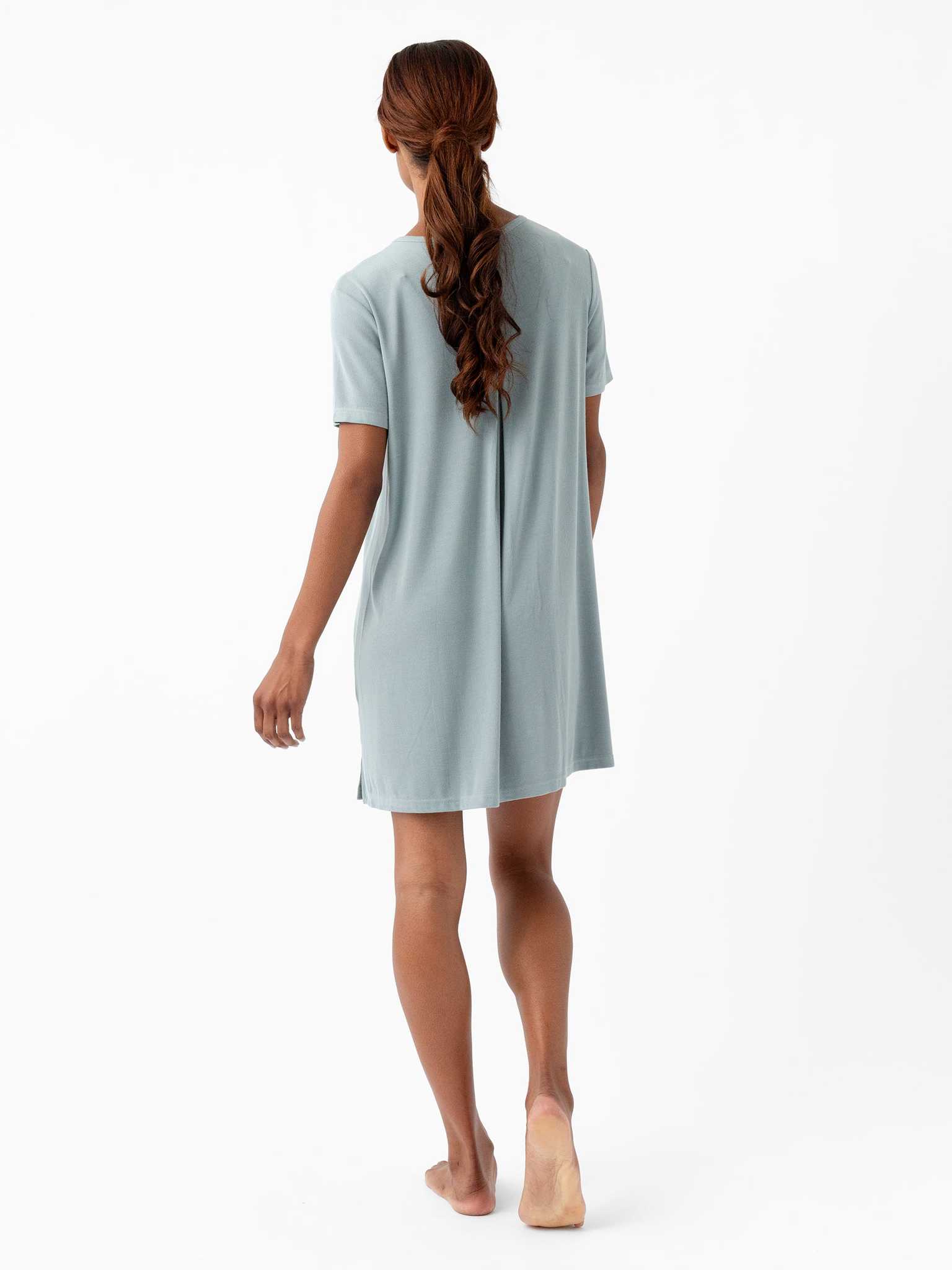 A person with long, wavy hair is shown from the back walking barefoot. They are wearing a Women's Bamboo Rib-Knit Boyfriend Sleep Dress by Cozy Earth in light blue, which has short sleeves and falls above the knees. The background is entirely white, emphasizing the subject and their attire. |Color:Smokey Blue