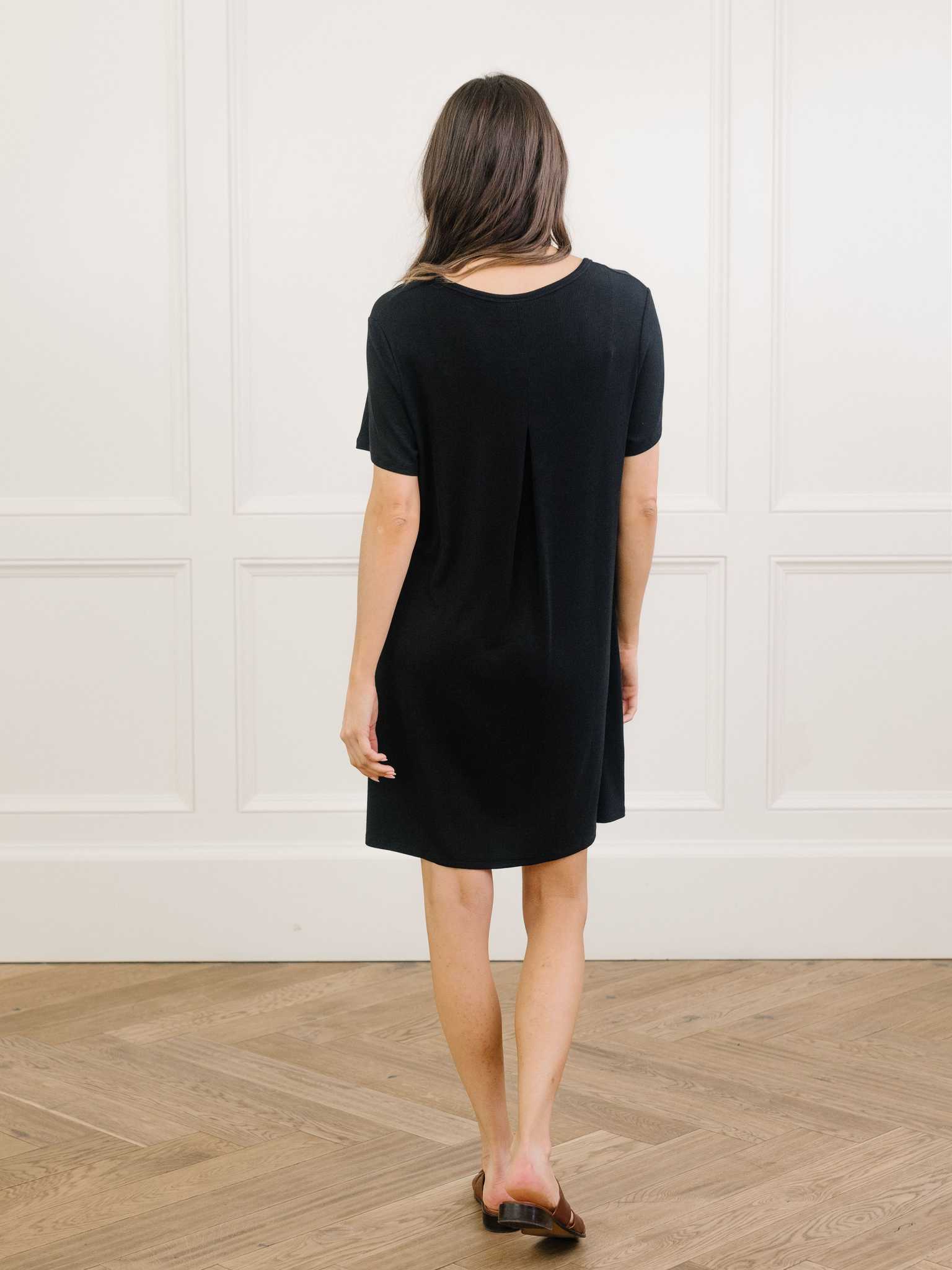 A woman facing backwards standing in a black Cozy Earth bamboo rib-knit boyfriend sleep dress with short sleeves, knee-length, in a lounge or bedroom setting. |Color:Black