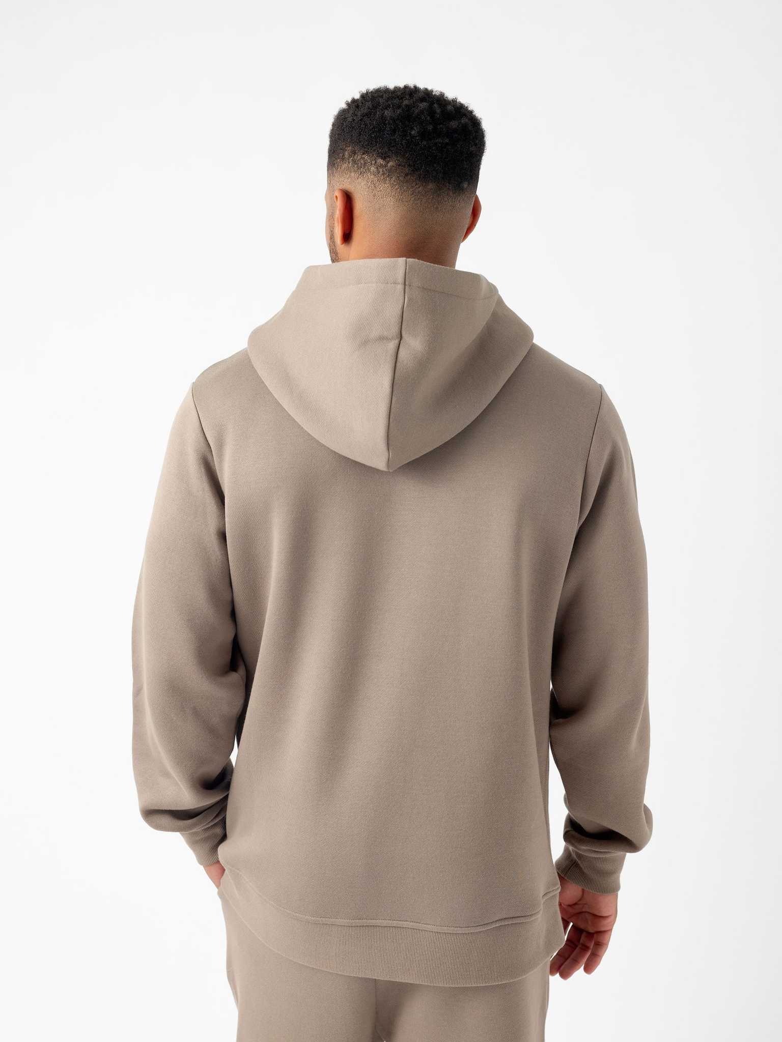 A person stands facing away from the camera, wearing a light brown Men's CityScape Hoodie and matching pants from Cozy Earth. The background is plain white, highlighting the outfit. The hoodie is plain with no visible designs or logos. |Color:Ebony