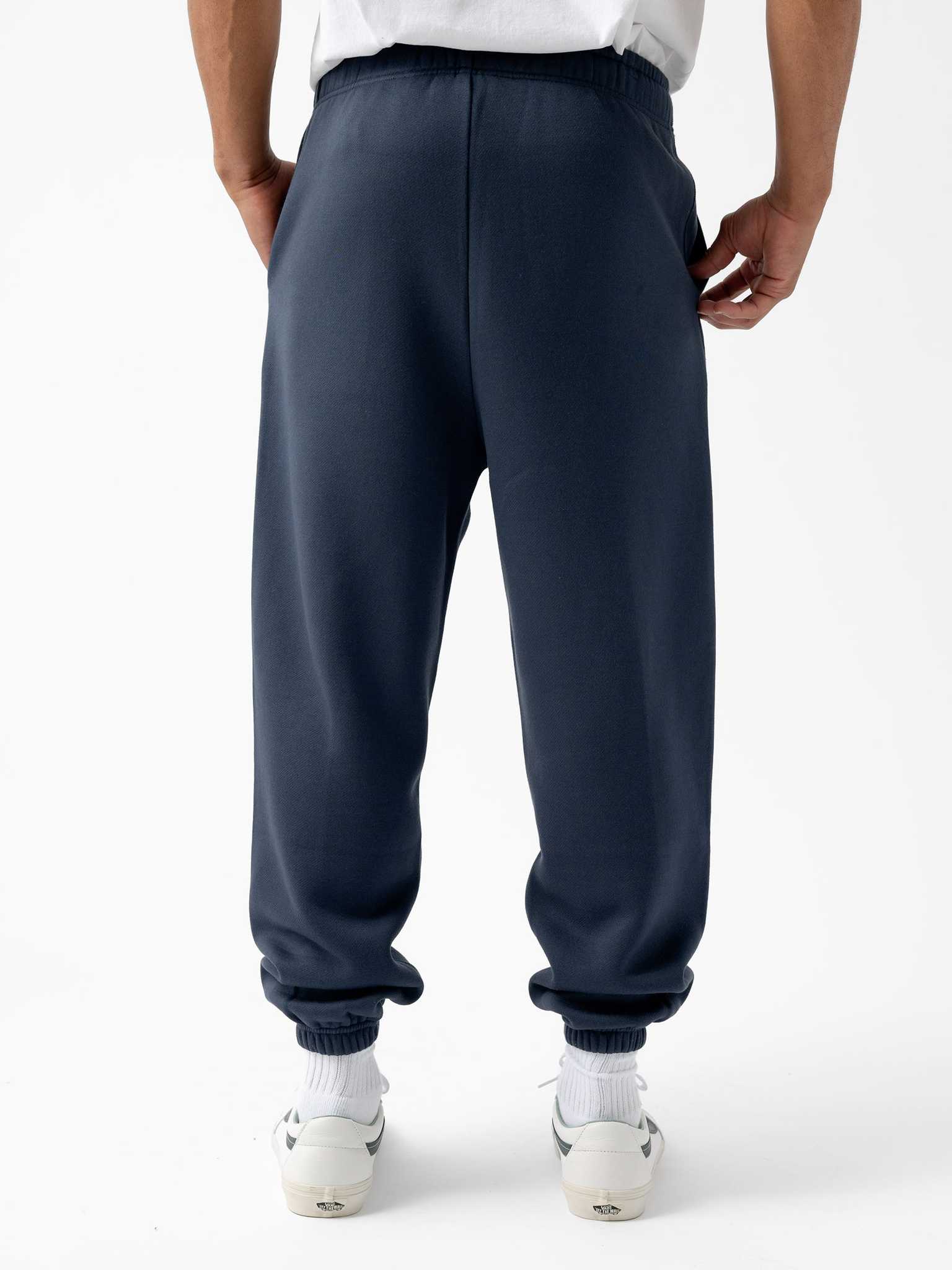 A person wearing the Men's CityScape Sweatpant by Cozy Earth in dark blue and white sneakers stands facing away, hands in pockets. The sweatpants feature elastic cuffs at the ankles. The background is plain white. |Color:Eclipse