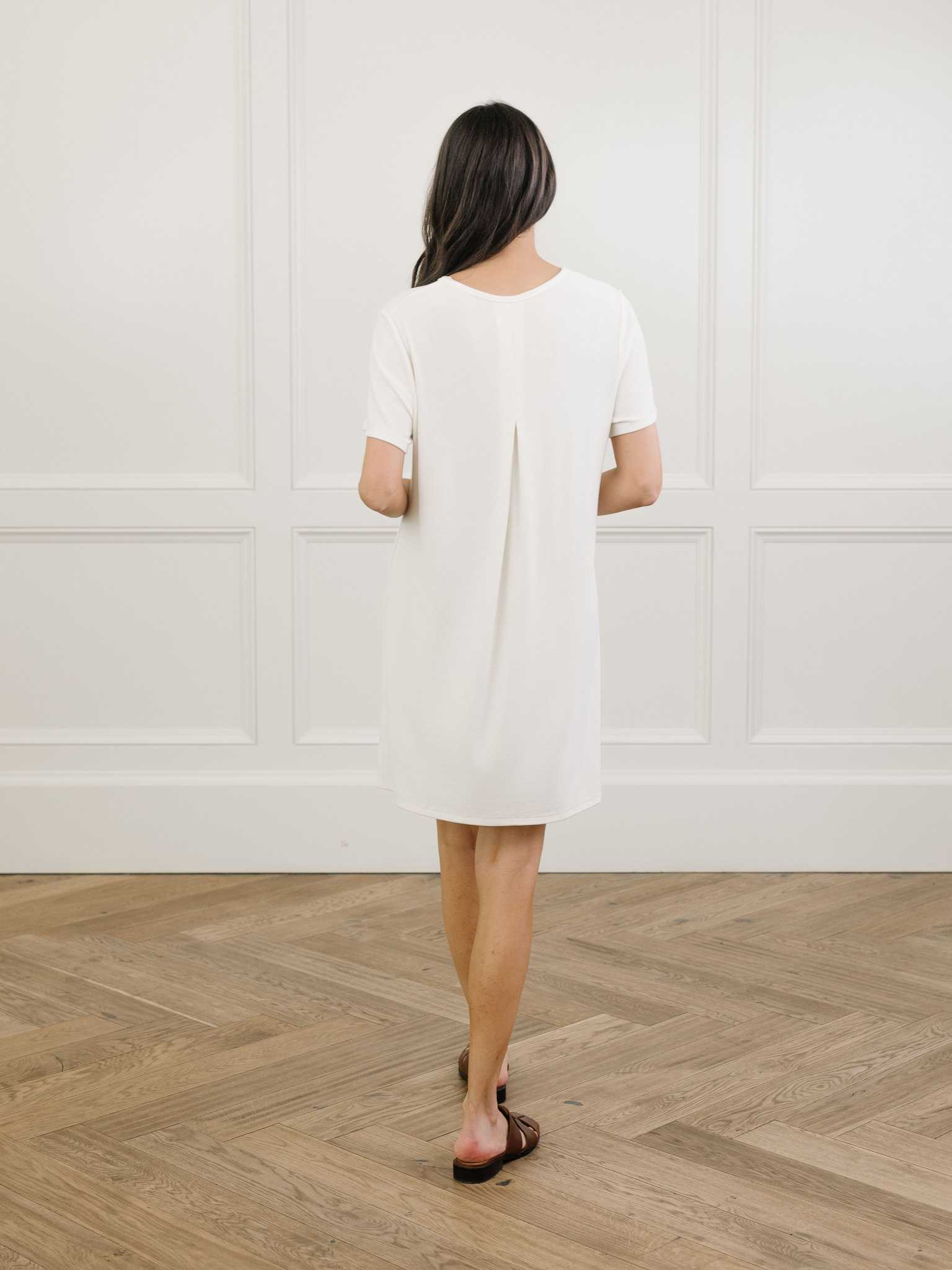 A facing backwards woman standing in a white Cozy Earth bamboo rib-knit boyfriend sleep dress with short sleeves, knee-length, in a lounge or bedroom setting. |Color:Ivory