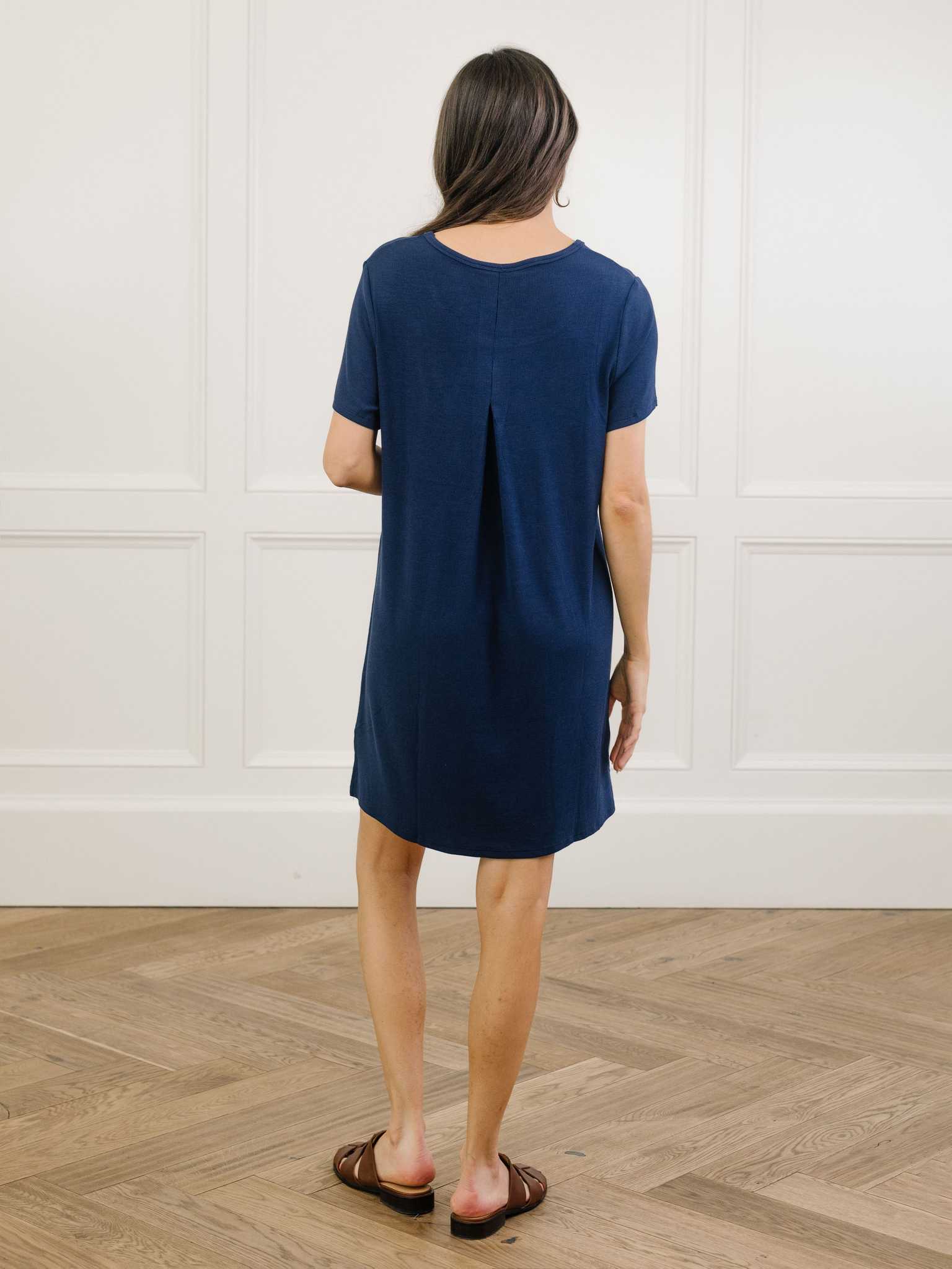 A woman facing backwards in a navy Cozy Earth bamboo rib-knit boyfriend sleep dress with short sleeves, knee-length, in a lounge or bedroom setting. |Color:Navy