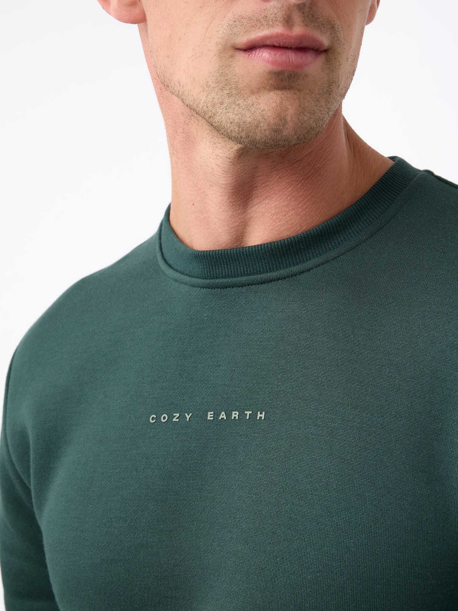 Close-up of a person wearing the Men's CityScape Crewneck, featuring the brand name "Cozy Earth" printed on the front. The focus is on the upper chest and neck area, against a plain white background. |Color:Spruce