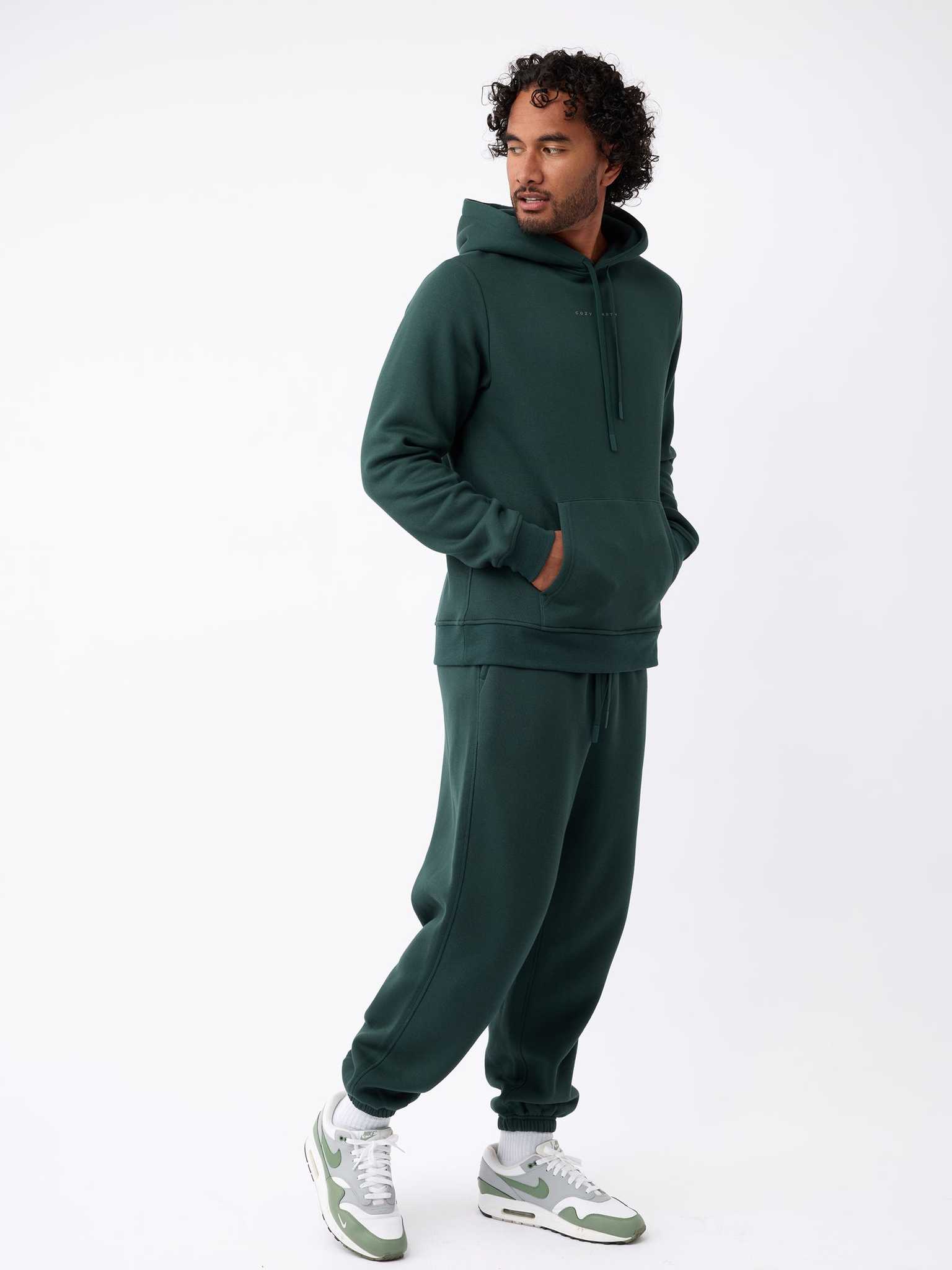 A man stands in a relaxed pose wearing a dark green hoodie paired with Cozy Earth's Men's CityScape Sweatpant. He has one hand in his pocket while looking to the side, and he complements his outfit with white and green sneakers against a white background. |Color:Spruce
