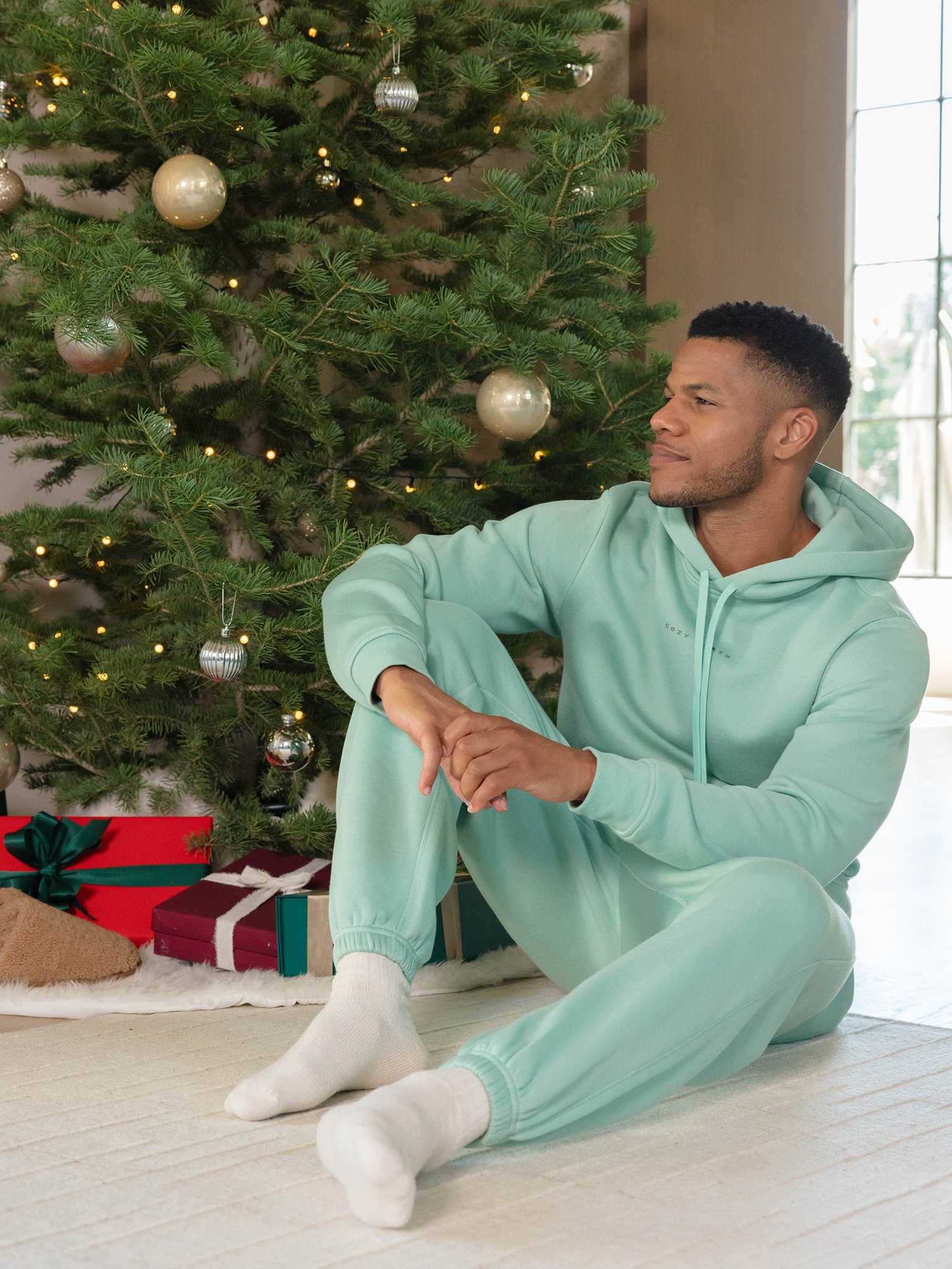 A person in a mint green Cozy Earth Men's CityScape Sweatpant sits on the floor near a decorated Christmas tree adorned with gold and silver ornaments. Wrapped gifts are nestled under the tree, and the room is softly lit. |Color:Wintergreen