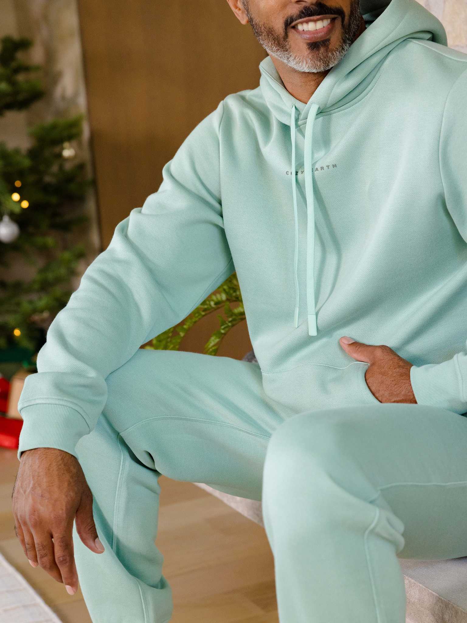 A person in a mint green Men's CityScape Hoodie and matching sweatpants from Cozy Earth sits casually. A Christmas tree with lights can be seen in the background, creating a festive atmosphere. |Color:Wintergreen