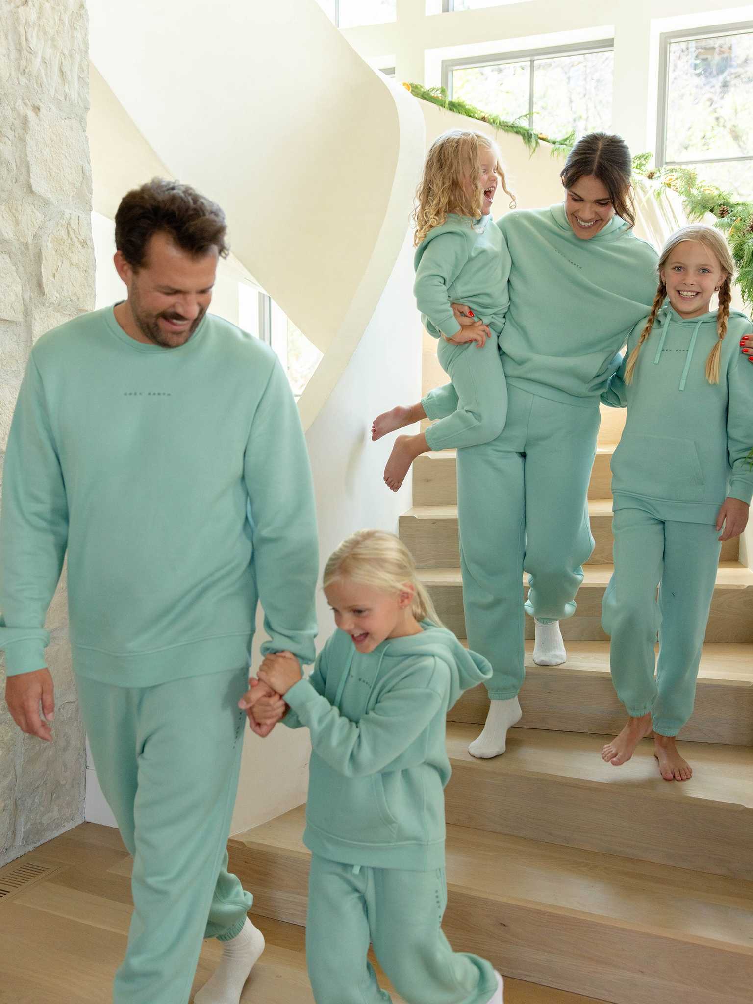 A family of five, clad in matching turquoise CityScape tracksuits by Cozy Earth, joyfully descends a modern staircase. The father, wearing the Men's CityScape Sweatpant, holds hands with one child at the front, while the mother carries another child and walks alongside their third child who is smiling brightly beside her. |Color:Wintergreen