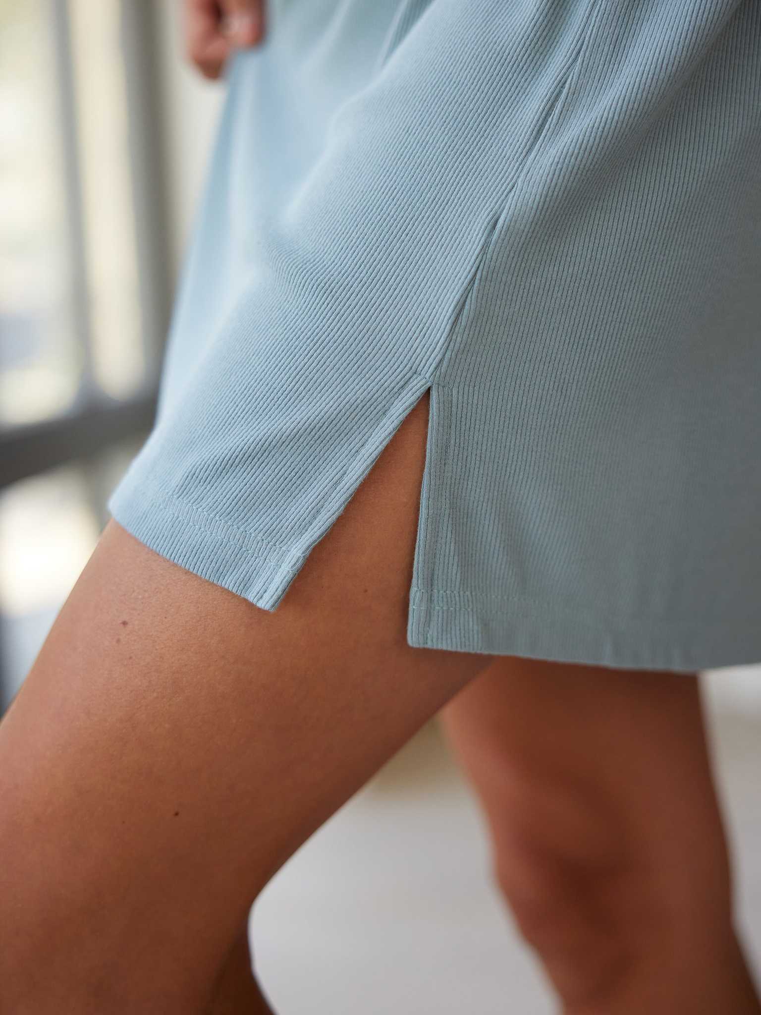 Close-up of the Women’s Bamboo Rib-Knit Boyfriend Sleep Dress by Cozy Earth in light blue, featuring a side slit that reveals part of a person's thigh and knee. The background is blurred. |Color:Smokey Blue